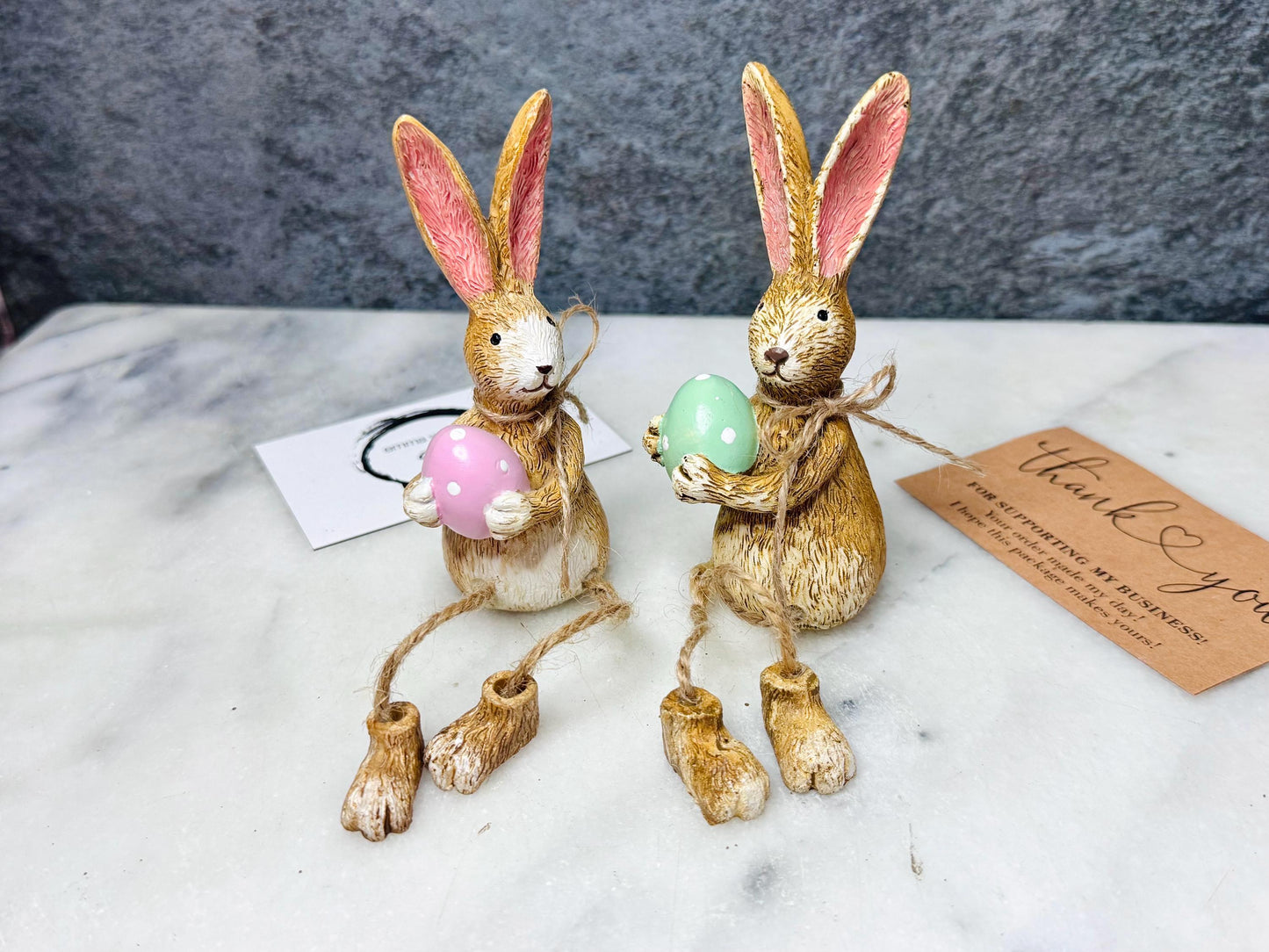 Hand-finished shelf sitting bunny ornament with dangling jute legs and polka dot egg. Available in pink or green. Perfect Easter rabbit shelf decoration, birthday gift, or home décor accessory. Cute animal figurine for rabbit lovers, Easter decorations, new home presents, or seasonal mantel styling.