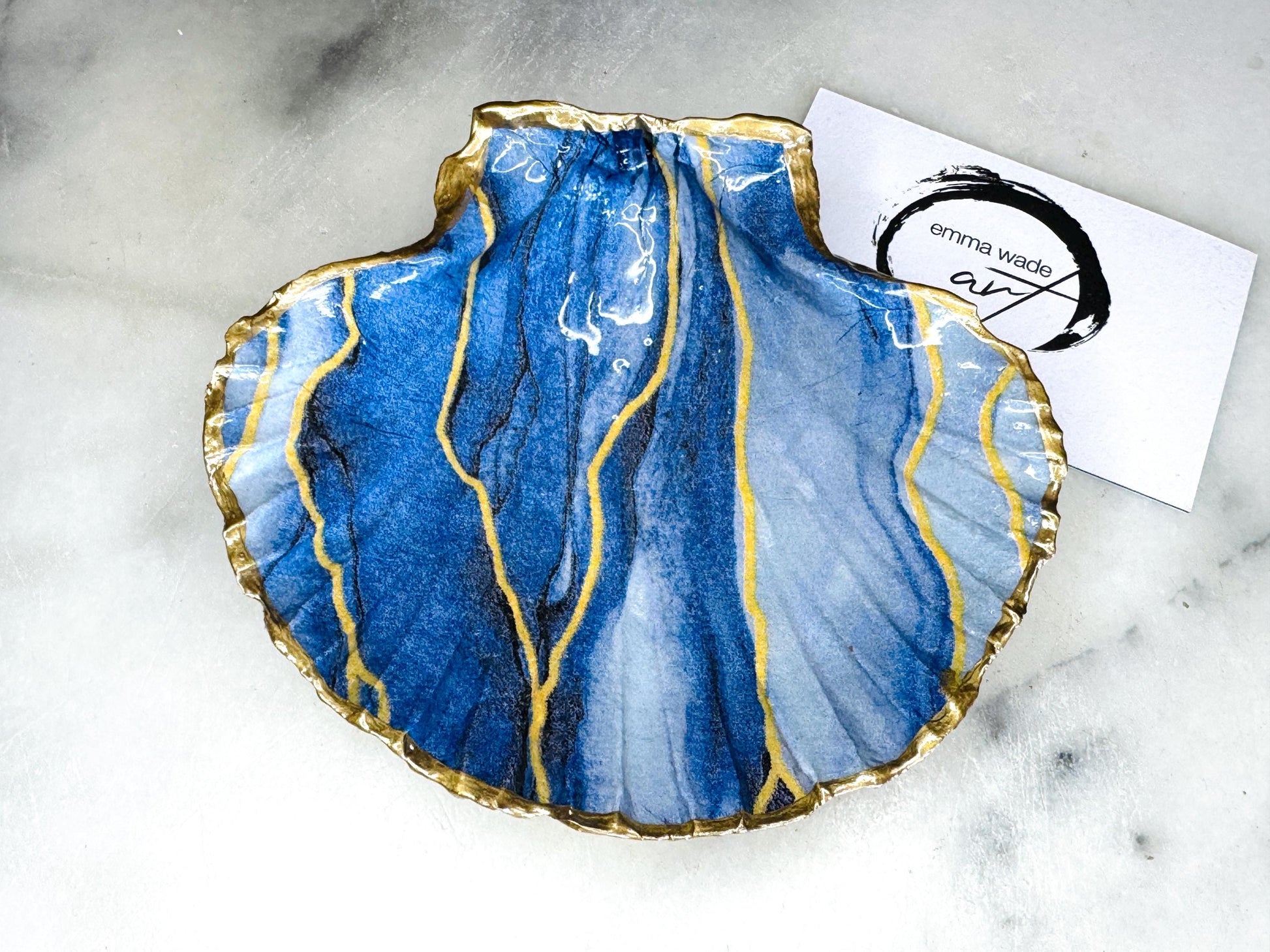 Hand painted blue waves scallop shell trinket dish with gold leaf accents and glossy resin finish, shown with optional gold driftwood display stand. Coastal jewellery and ring dish crafted from a natural Dorset scallop shell, featuring flowing blue wave artwork. Decorative shell bowl photographed on a light background, showing the natural shell shape and gilded edges, ideal for holding rings, earrings, trinkets or small treasures.