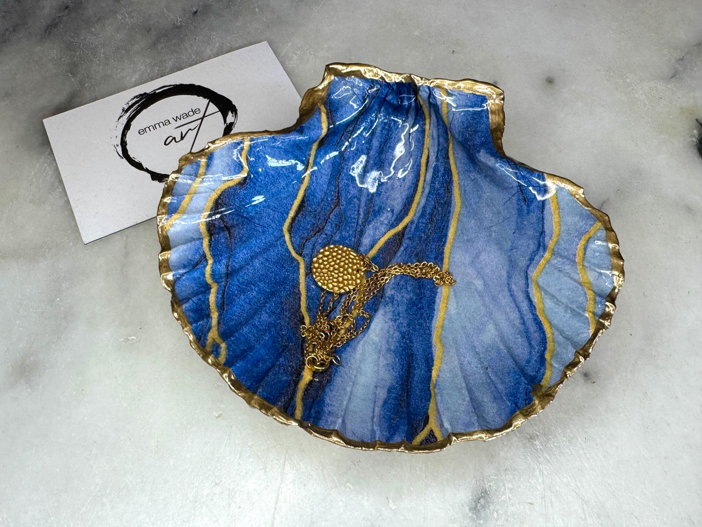 Hand painted blue waves scallop shell trinket dish with gold leaf accents and glossy resin finish, shown with optional gold driftwood display stand. Coastal jewellery and ring dish crafted from a natural Dorset scallop shell, featuring flowing blue wave artwork. Decorative shell bowl photographed on a light background, showing the natural shell shape and gilded edges, ideal for holding rings, earrings, trinkets or small treasures.
