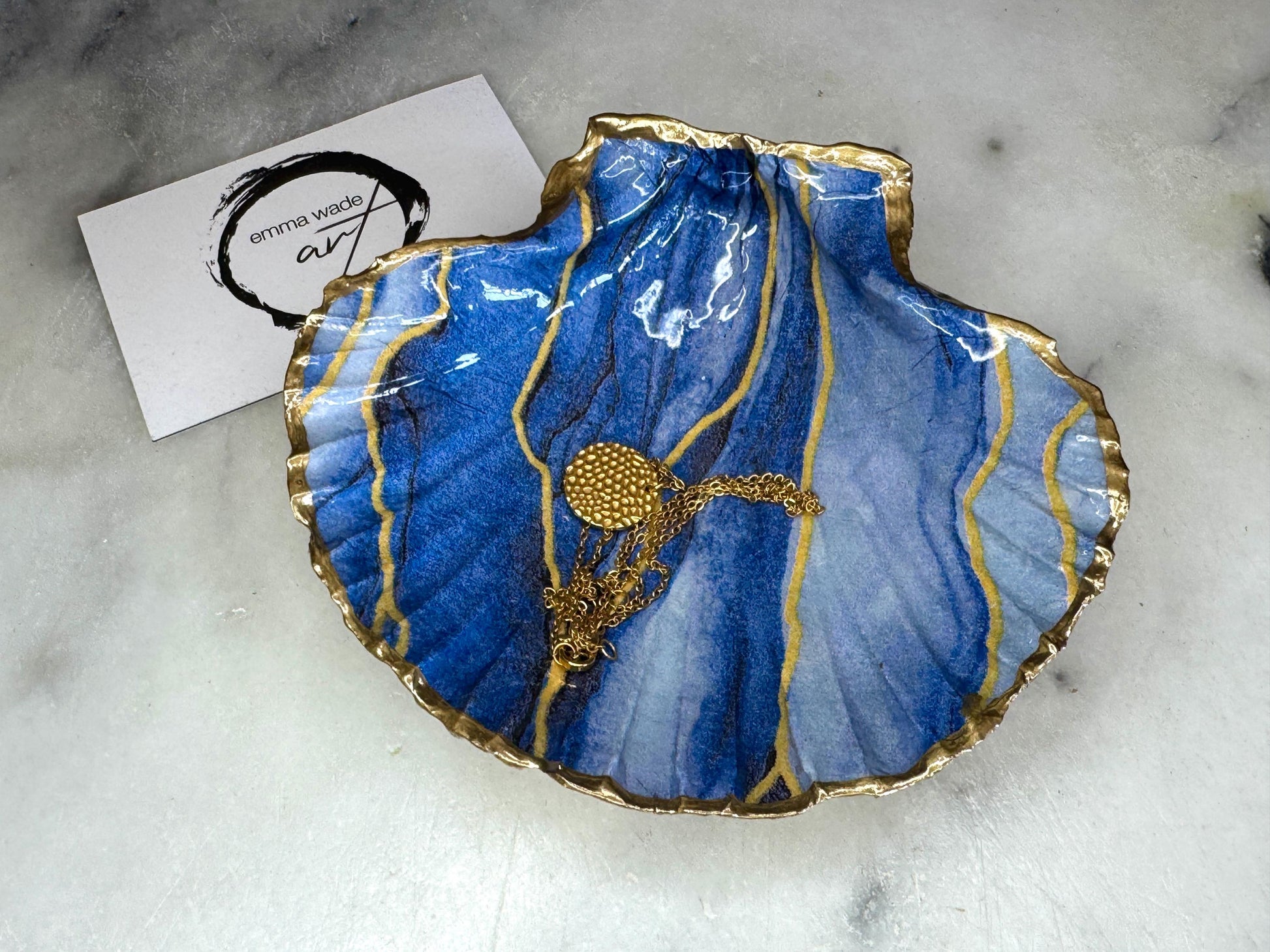 Hand painted blue waves scallop shell trinket dish with gold leaf accents and glossy resin finish, shown with optional gold driftwood display stand. Coastal jewellery and ring dish crafted from a natural Dorset scallop shell, featuring flowing blue wave artwork. Decorative shell bowl photographed on a light background, showing the natural shell shape and gilded edges, ideal for holding rings, earrings, trinkets or small treasures.