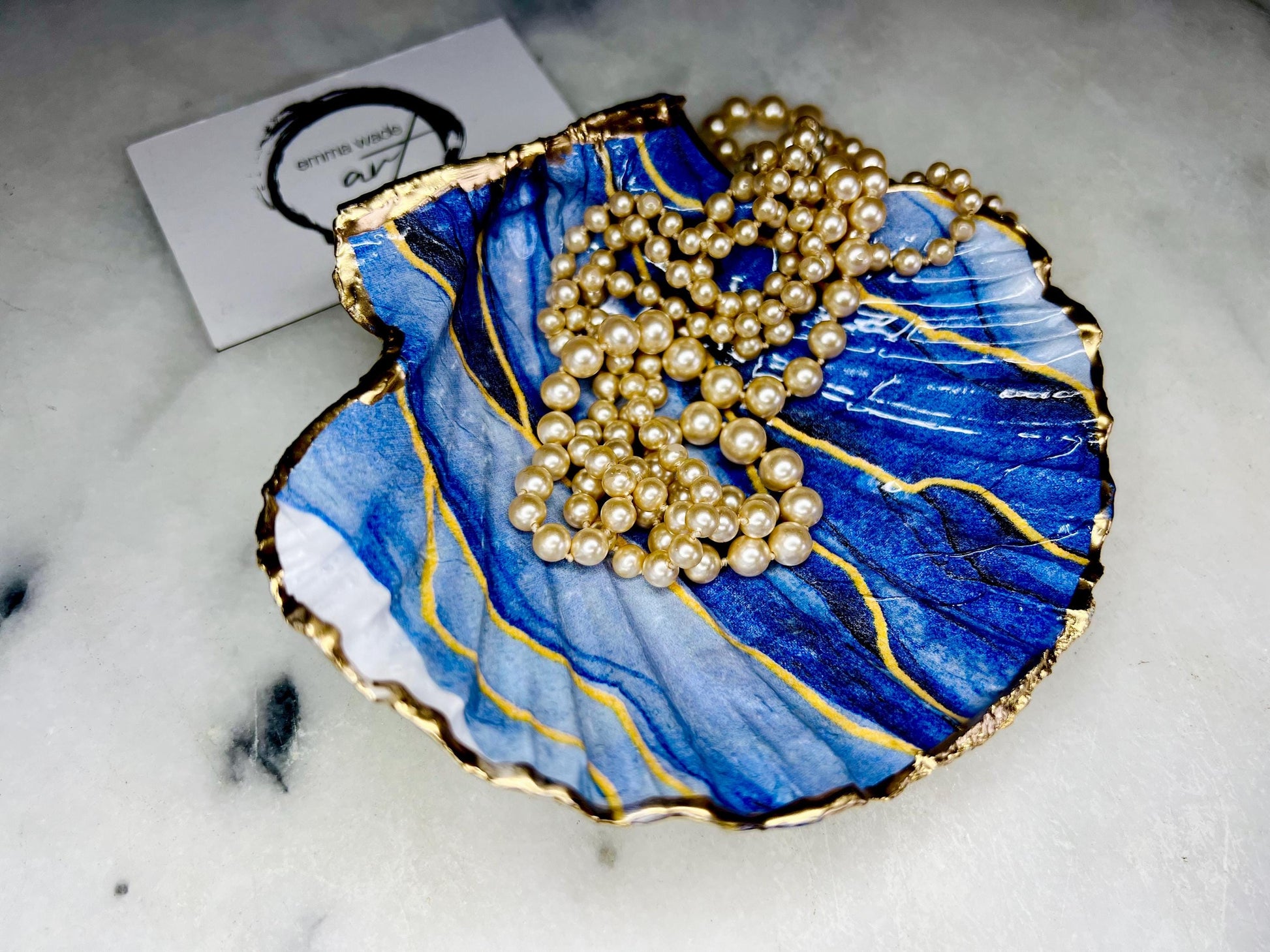 Hand painted blue waves scallop shell trinket dish with gold leaf accents and glossy resin finish, shown with optional gold driftwood display stand. Coastal jewellery and ring dish crafted from a natural Dorset scallop shell, featuring flowing blue wave artwork. Decorative shell bowl photographed on a light background, showing the natural shell shape and gilded edges, ideal for holding rings, earrings, trinkets or small treasures.