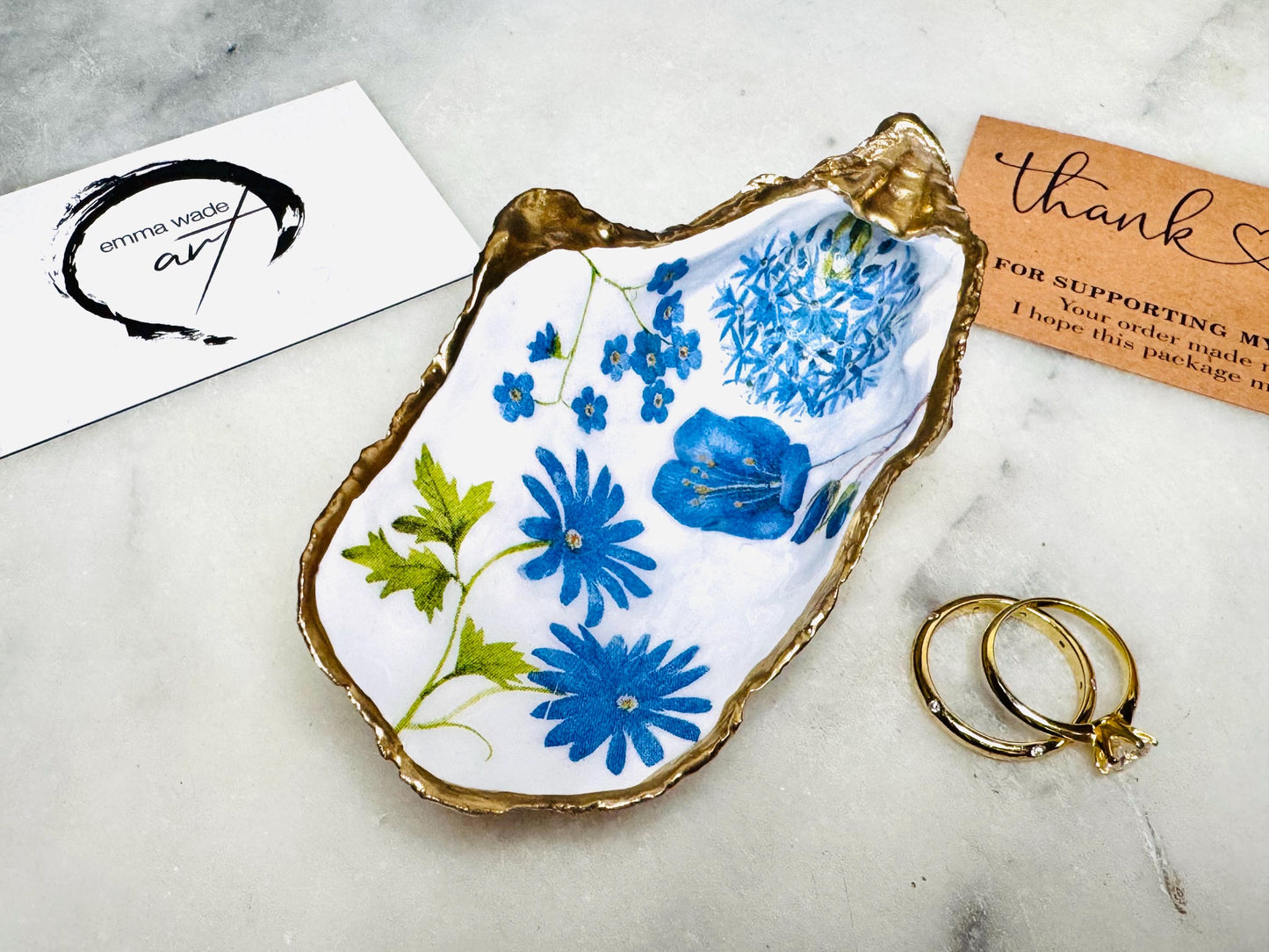 Handmade Dorset oyster shell trinket dish featuring delicate blue floral artwork including bluebells and forget-me-nots, finished with hand-applied gold leaf edging and glossy resin. Coastal-inspired jewellery dish and ring holder available in multiple sizes including XL oyster shells. Elegant handmade home décor gift ideal for birthdays, bridesmaids, Mother’s Day, weddings, and coastal interiors.