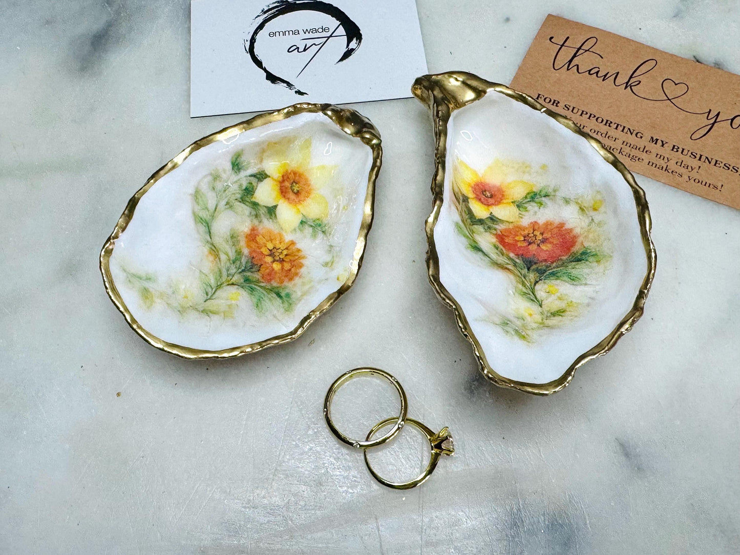 Handmade Dorset oyster shell trinket dish with yellow and orange spring flowers, available with green foliage or stylised blue-toned foliage, finished with a gold leaf rim and glossy resin. Features an original Emma Wade Art design created exclusively for this collection. Shown holding rings on a light background, highlighting the natural shell shape and handcrafted coastal style, ideal as a jewellery dish or keepsake gift.