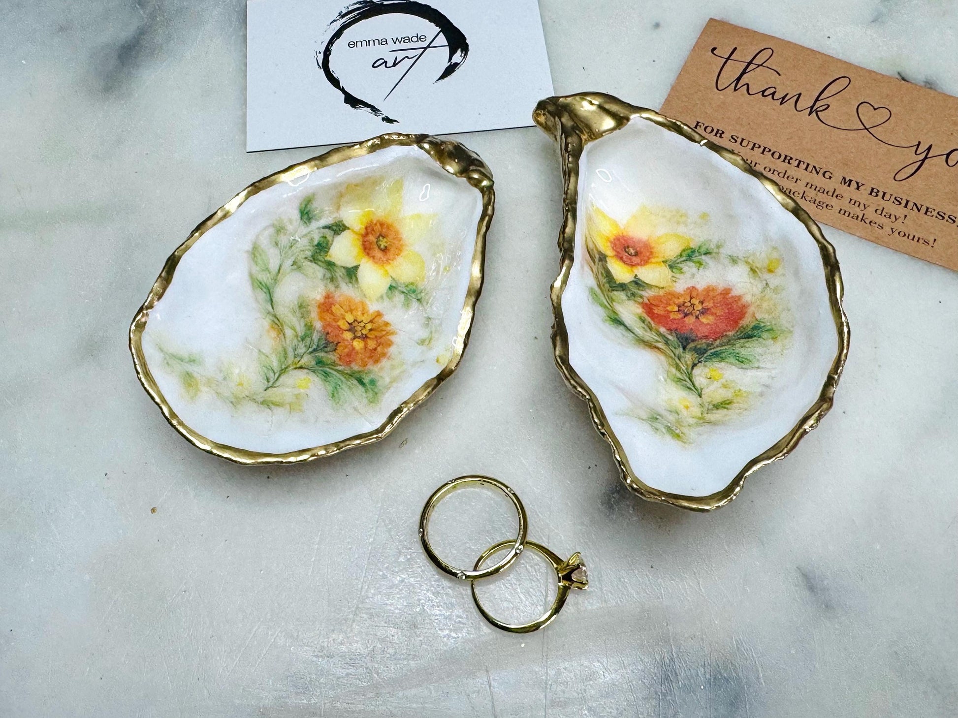 Handmade Dorset oyster shell trinket dish with yellow and orange spring flowers, available with green foliage or stylised blue-toned foliage, finished with a gold leaf rim and glossy resin. Features an original Emma Wade Art design created exclusively for this collection. Shown holding rings on a light background, highlighting the natural shell shape and handcrafted coastal style, ideal as a jewellery dish or keepsake gift.