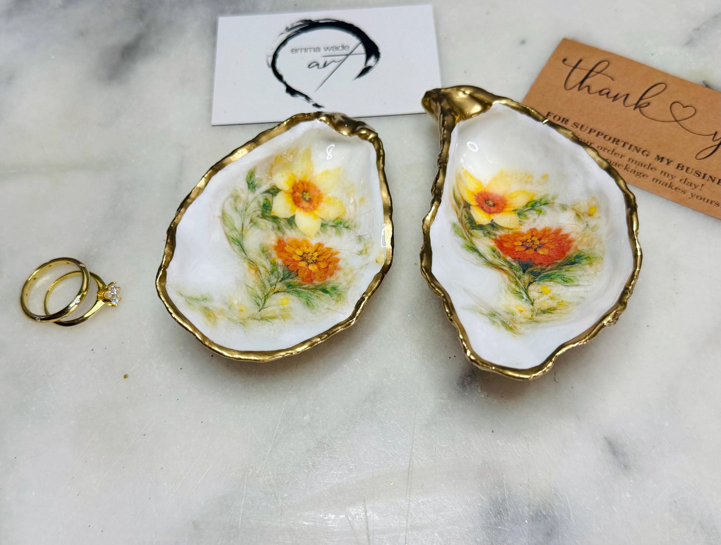 Handmade Dorset oyster shell trinket dish with yellow and orange spring flowers, available with green foliage or stylised blue-toned foliage, finished with a gold leaf rim and glossy resin. Features an original Emma Wade Art design created exclusively for this collection. Shown holding rings on a light background, highlighting the natural shell shape and handcrafted coastal style, ideal as a jewellery dish or keepsake gift.