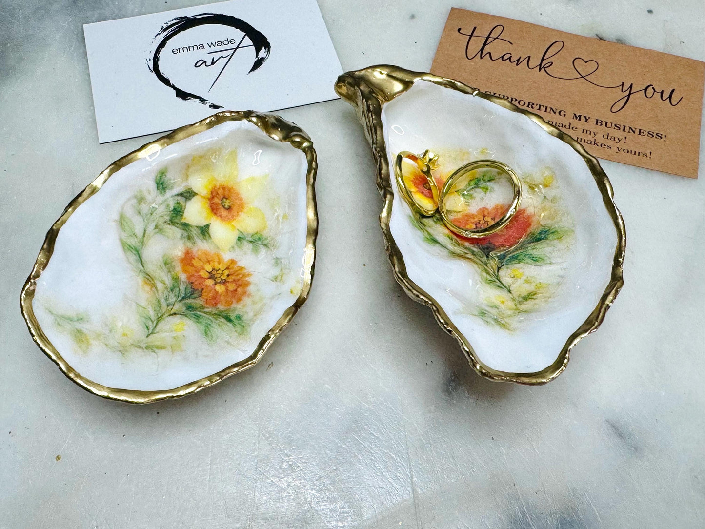 Handmade Dorset oyster shell trinket dish with yellow and orange spring flowers, available with green foliage or stylised blue-toned foliage, finished with a gold leaf rim and glossy resin. Features an original Emma Wade Art design created exclusively for this collection. Shown holding rings on a light background, highlighting the natural shell shape and handcrafted coastal style, ideal as a jewellery dish or keepsake gift.
