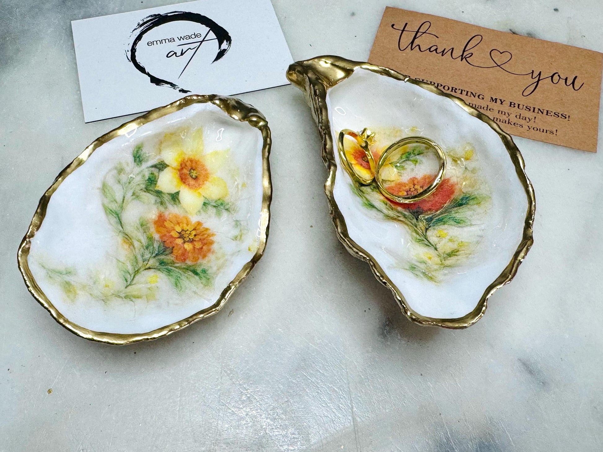 Handmade Dorset oyster shell trinket dish with yellow and orange spring flowers, available with green foliage or stylised blue-toned foliage, finished with a gold leaf rim and glossy resin. Features an original Emma Wade Art design created exclusively for this collection. Shown holding rings on a light background, highlighting the natural shell shape and handcrafted coastal style, ideal as a jewellery dish or keepsake gift.