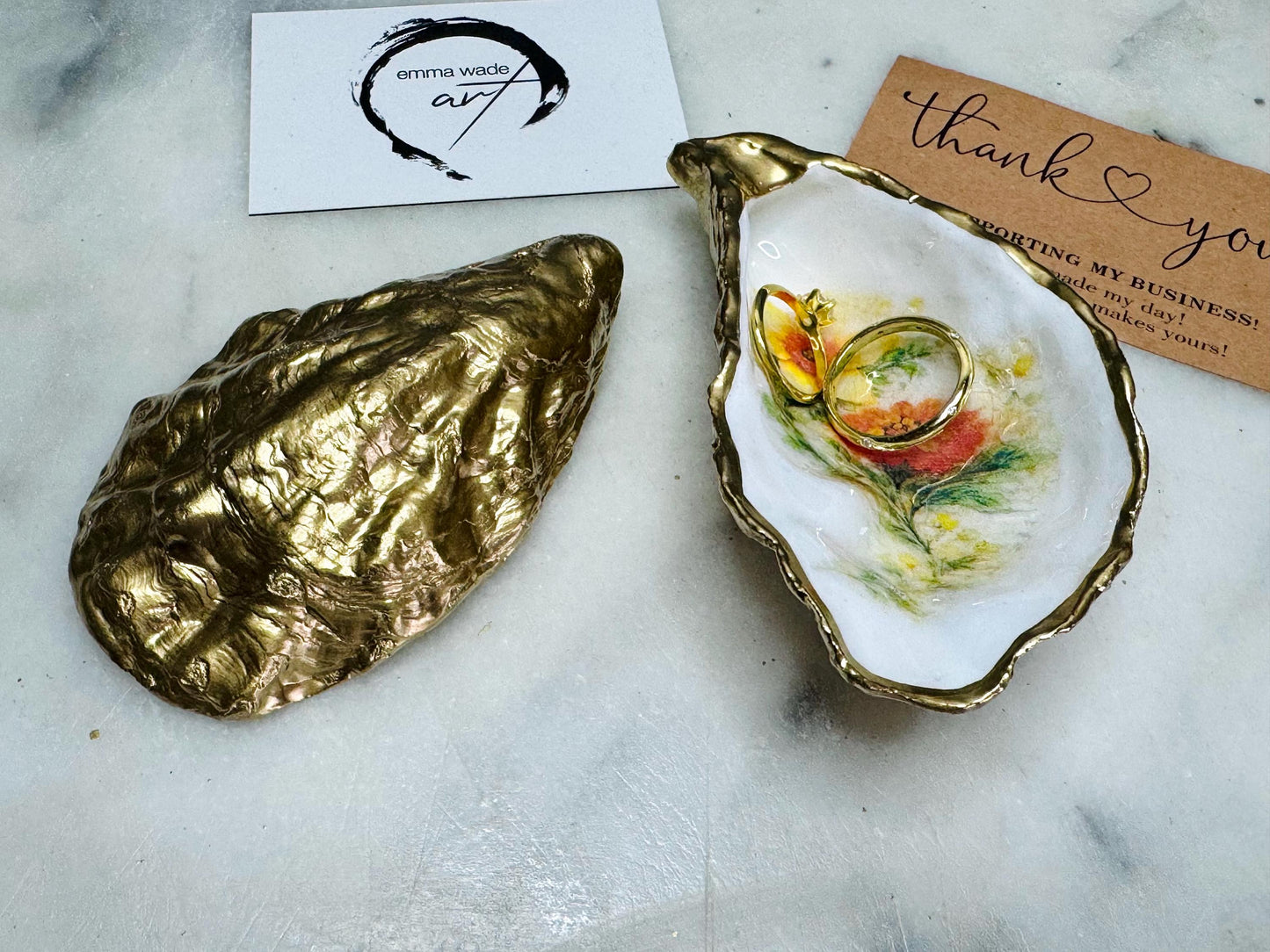 Handmade Dorset oyster shell trinket dish with yellow and orange spring flowers, available with green foliage or stylised blue-toned foliage, finished with a gold leaf rim and glossy resin. Features an original Emma Wade Art design created exclusively for this collection. Shown holding rings on a light background, highlighting the natural shell shape and handcrafted coastal style, ideal as a jewellery dish or keepsake gift.