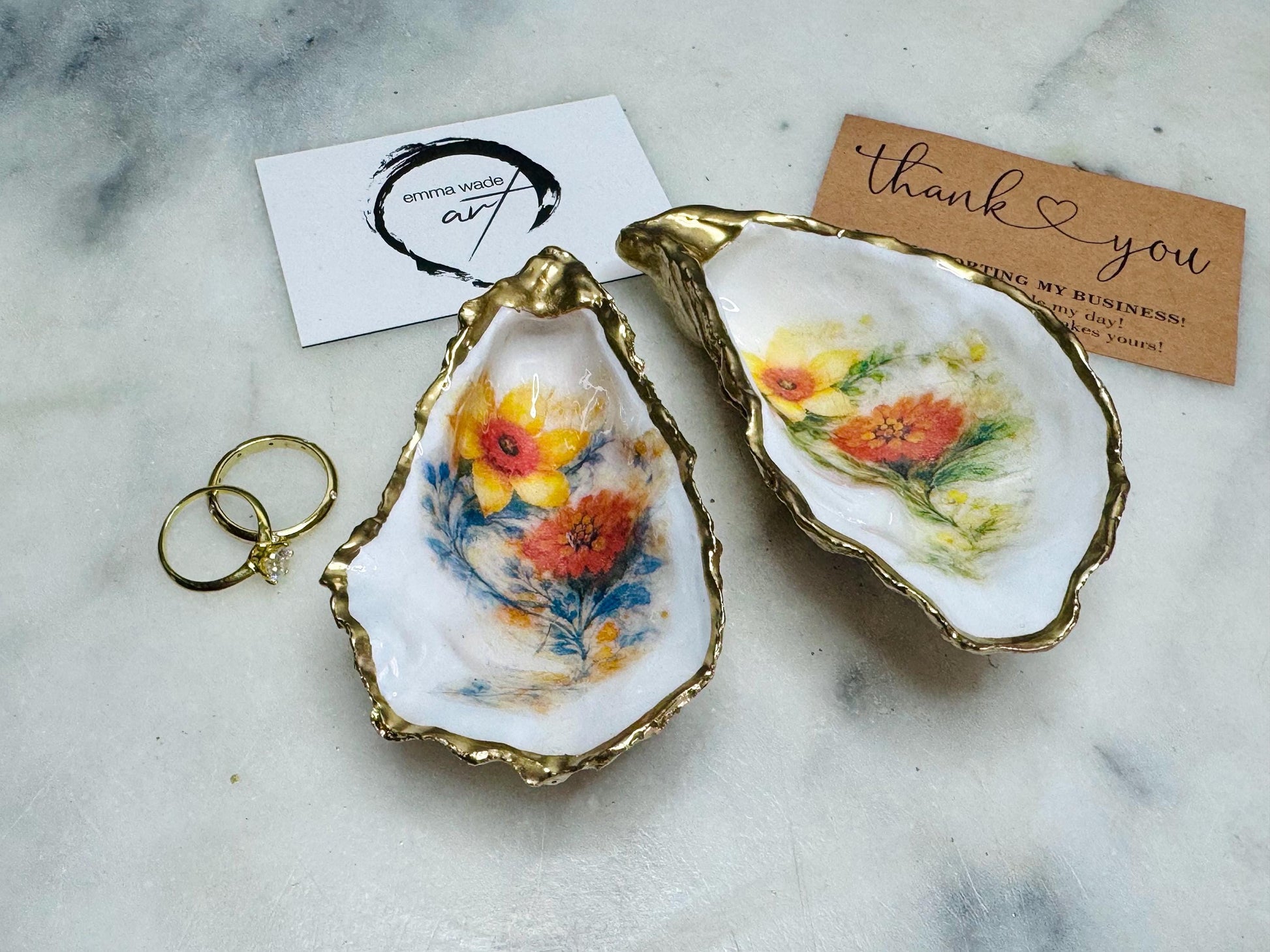 Handmade Dorset oyster shell trinket dish with yellow and orange spring flowers, available with green foliage or stylised blue-toned foliage, finished with a gold leaf rim and glossy resin. Features an original Emma Wade Art design created exclusively for this collection. Shown holding rings on a light background, highlighting the natural shell shape and handcrafted coastal style, ideal as a jewellery dish or keepsake gift.