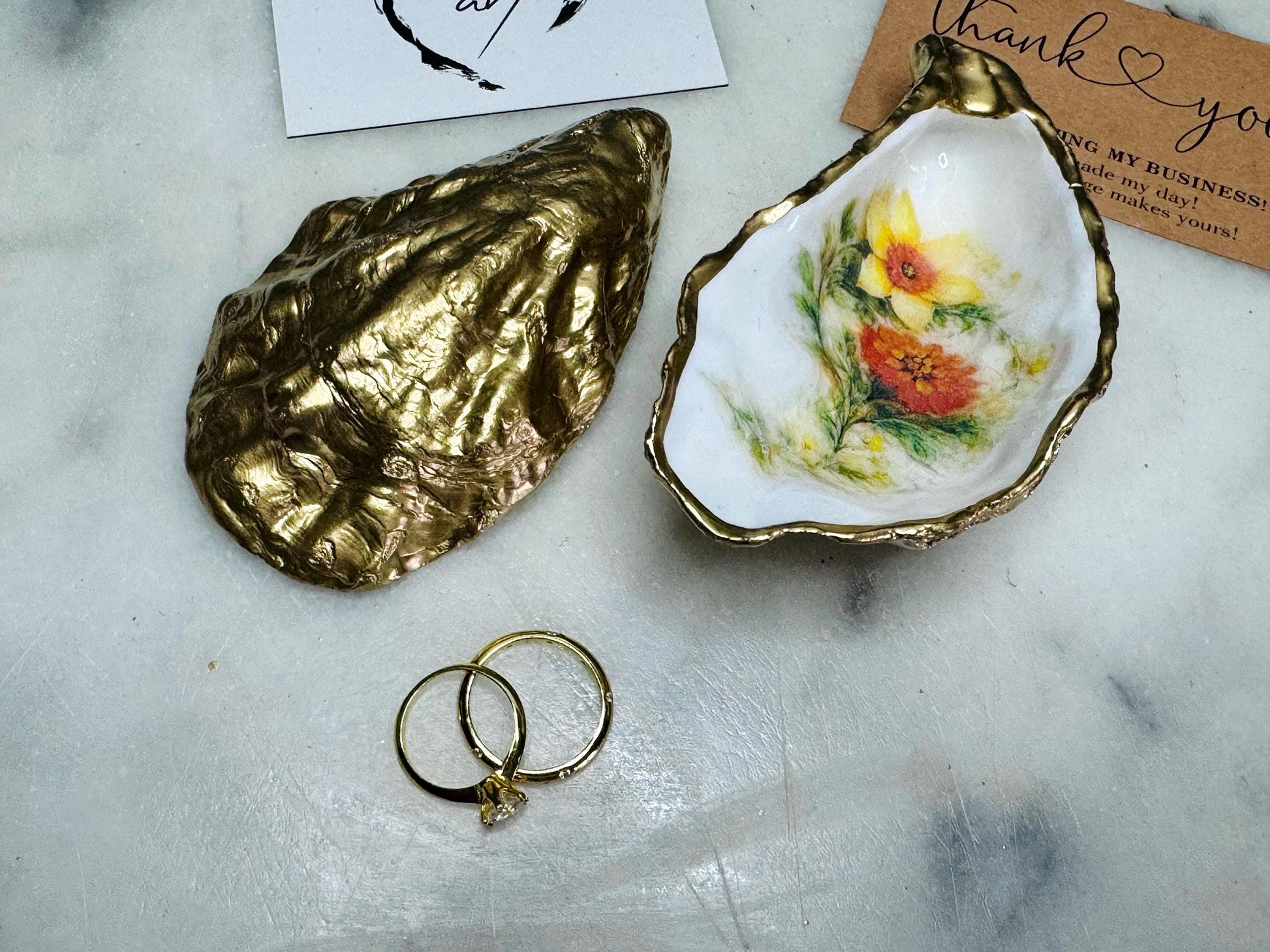 Handmade Dorset oyster shell trinket dish with yellow and orange spring flowers, available with green foliage or stylised blue-toned foliage, finished with a gold leaf rim and glossy resin. Features an original Emma Wade Art design created exclusively for this collection. Shown holding rings on a light background, highlighting the natural shell shape and handcrafted coastal style, ideal as a jewellery dish or keepsake gift.