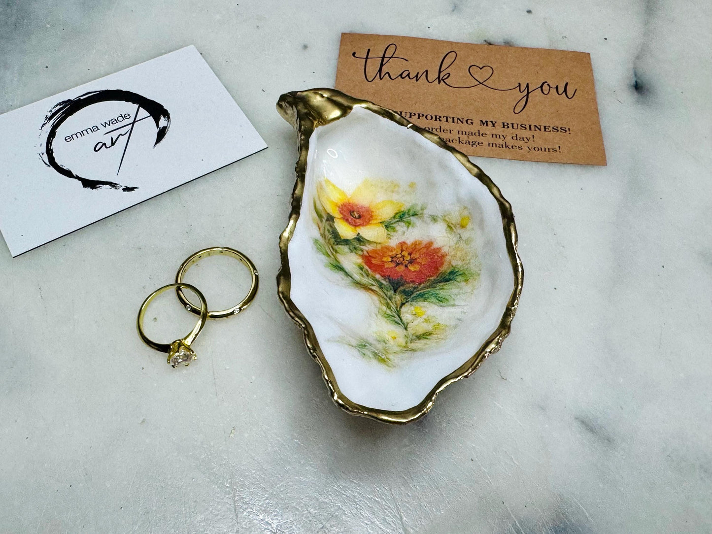 Handmade Dorset oyster shell trinket dish with yellow and orange spring flowers, available with green foliage or stylised blue-toned foliage, finished with a gold leaf rim and glossy resin. Features an original Emma Wade Art design created exclusively for this collection. Shown holding rings on a light background, highlighting the natural shell shape and handcrafted coastal style, ideal as a jewellery dish or keepsake gift.