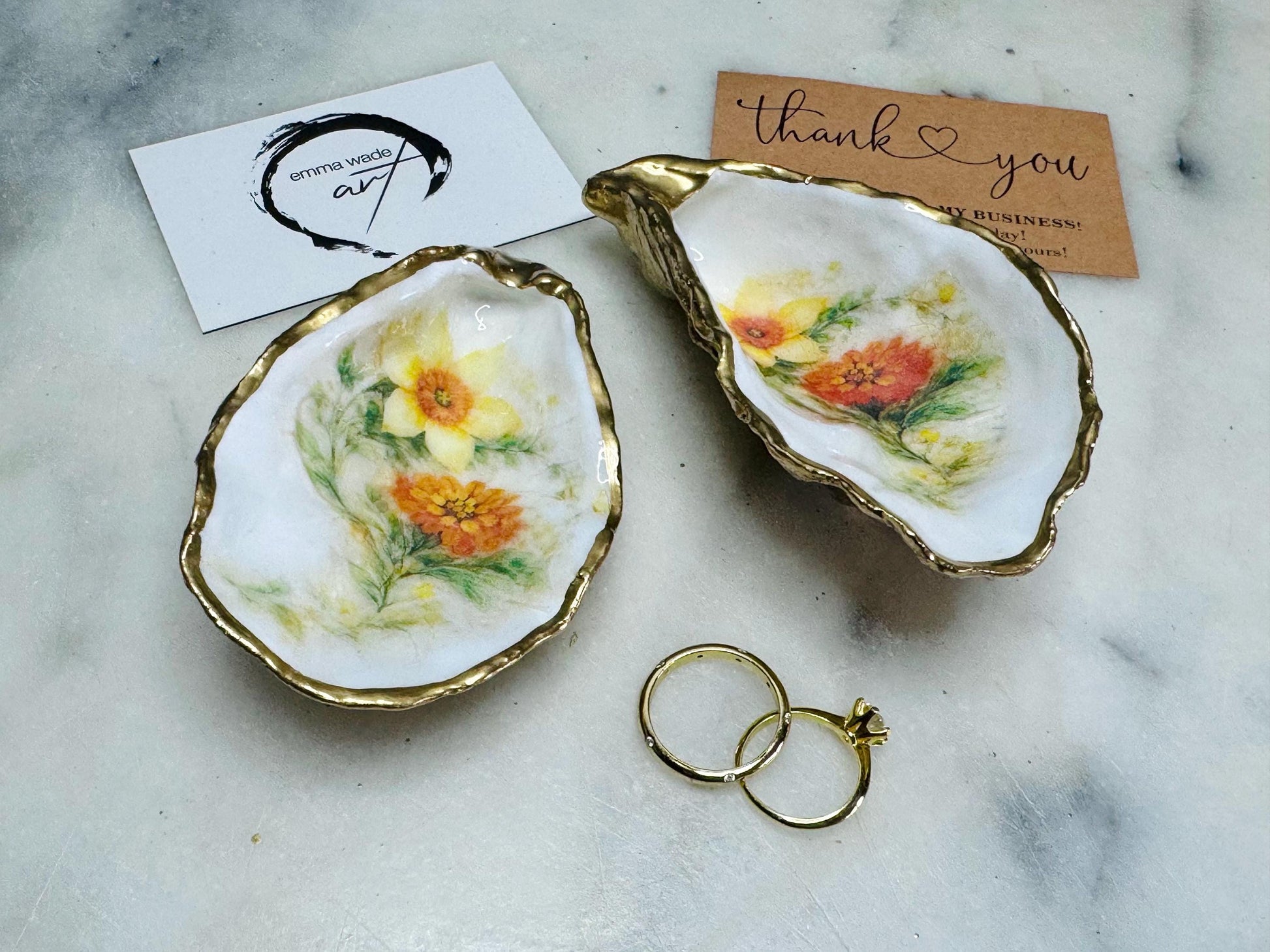 Handmade Dorset oyster shell trinket dish with yellow and orange spring flowers, available with green foliage or stylised blue-toned foliage, finished with a gold leaf rim and glossy resin. Features an original Emma Wade Art design created exclusively for this collection. Shown holding rings on a light background, highlighting the natural shell shape and handcrafted coastal style, ideal as a jewellery dish or keepsake gift.