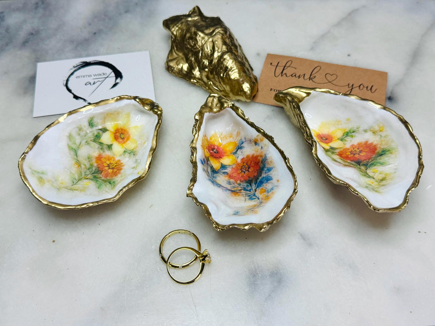 Handmade Dorset oyster shell trinket dish with yellow and orange spring flowers, available with green foliage or stylised blue-toned foliage, finished with a gold leaf rim and glossy resin. Features an original Emma Wade Art design created exclusively for this collection. Shown holding rings on a light background, highlighting the natural shell shape and handcrafted coastal style, ideal as a jewellery dish or keepsake gift.