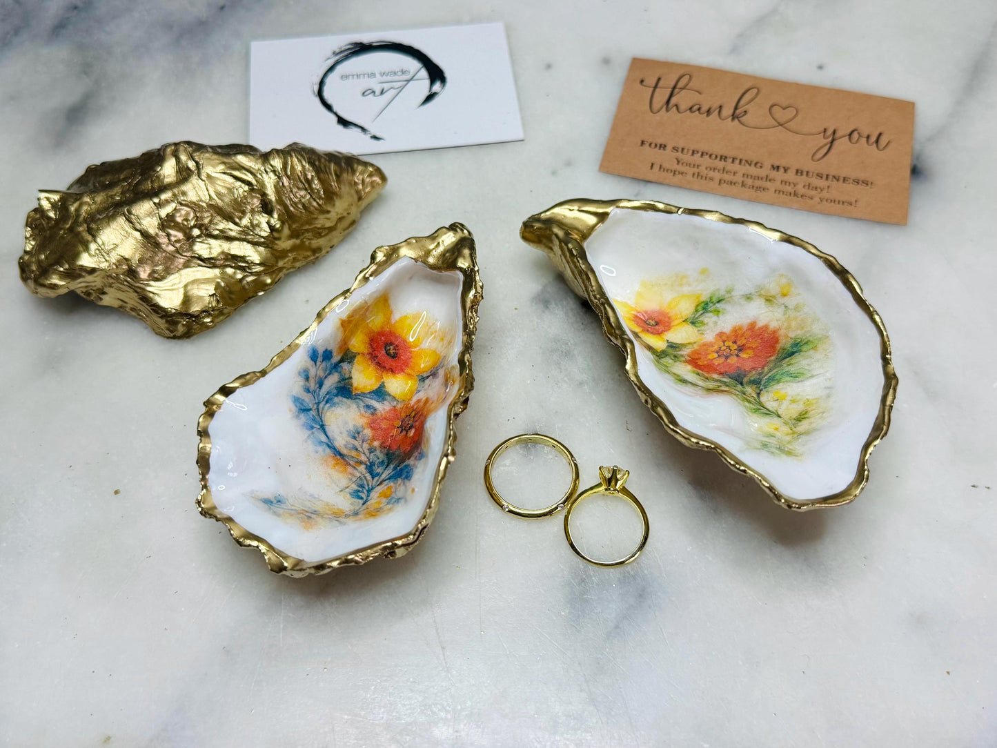 Handmade Dorset oyster shell trinket dish with yellow and orange spring flowers, available with green foliage or stylised blue-toned foliage, finished with a gold leaf rim and glossy resin. Features an original Emma Wade Art design created exclusively for this collection. Shown holding rings on a light background, highlighting the natural shell shape and handcrafted coastal style, ideal as a jewellery dish or keepsake gift.