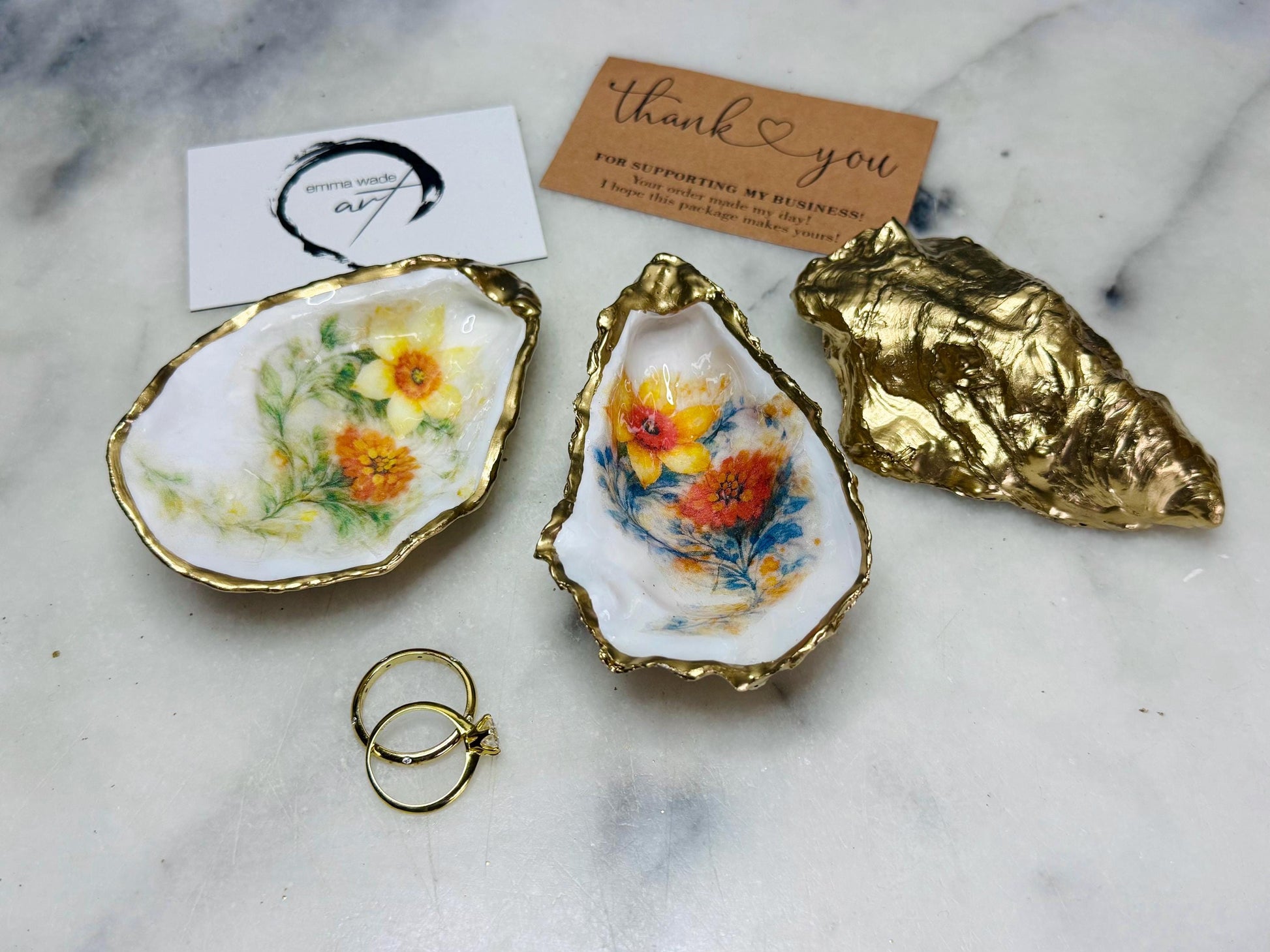 Handmade Dorset oyster shell trinket dish with yellow and orange spring flowers, available with green foliage or stylised blue-toned foliage, finished with a gold leaf rim and glossy resin. Features an original Emma Wade Art design created exclusively for this collection. Shown holding rings on a light background, highlighting the natural shell shape and handcrafted coastal style, ideal as a jewellery dish or keepsake gift.