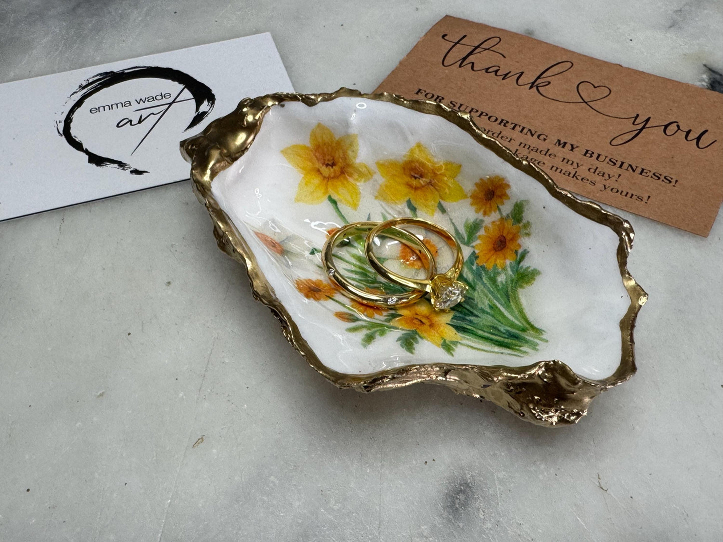 Handmade Dorset oyster shell trinket dish with yellow and orange spring flowers, Daffodils and Marigolds, available in two designs, finished with a gold leaf rim and glossy resin. Features an original Emma Wade Art design created exclusively for this collection. Shown holding rings on a light background, highlighting the natural shell shape and handcrafted coastal style, ideal as a jewellery dish or keepsake gift.