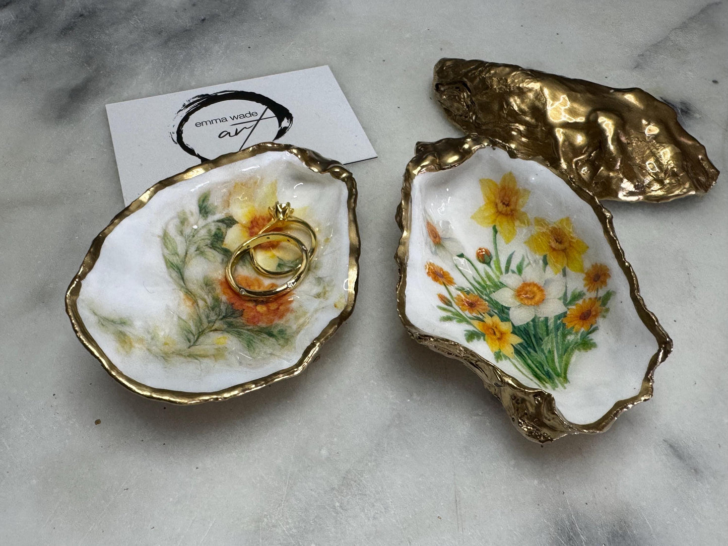 Handmade Dorset oyster shell trinket dish with yellow and orange spring flowers, Daffodils and Marigolds, available in two designs, finished with a gold leaf rim and glossy resin. Features an original Emma Wade Art design created exclusively for this collection. Shown holding rings on a light background, highlighting the natural shell shape and handcrafted coastal style, ideal as a jewellery dish or keepsake gift.