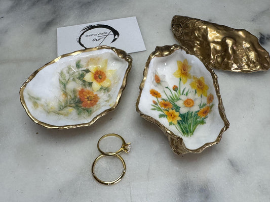 Handmade Dorset oyster shell trinket dish with yellow and orange spring flowers, Daffodils and Marigolds, available in two designs, finished with a gold leaf rim and glossy resin. Features an original Emma Wade Art design created exclusively for this collection. Shown holding rings on a light background, highlighting the natural shell shape and handcrafted coastal style, ideal as a jewellery dish or keepsake gift.