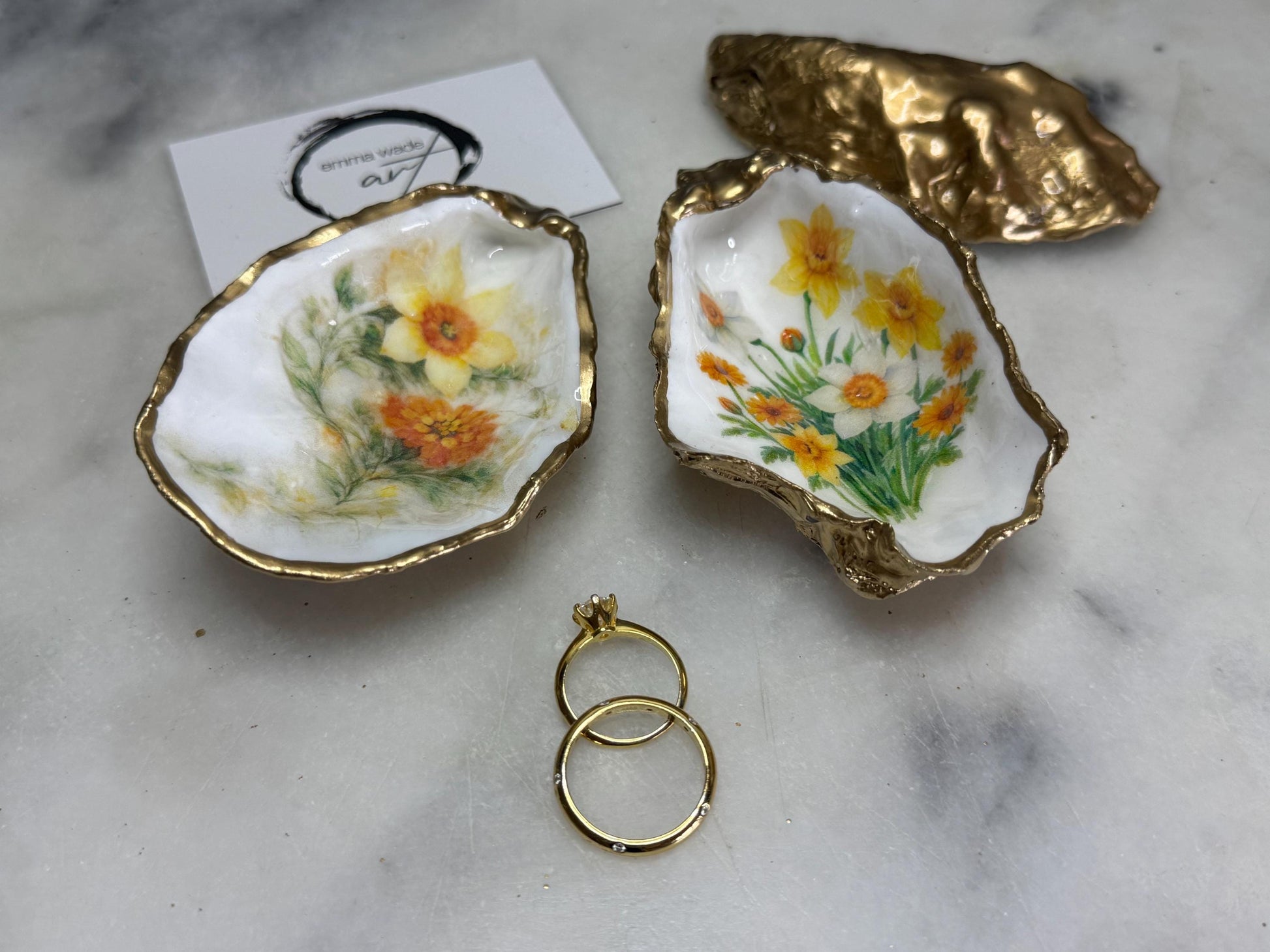 Handmade Dorset oyster shell trinket dish with yellow and orange spring flowers, Daffodils and Marigolds, available in two designs, finished with a gold leaf rim and glossy resin. Features an original Emma Wade Art design created exclusively for this collection. Shown holding rings on a light background, highlighting the natural shell shape and handcrafted coastal style, ideal as a jewellery dish or keepsake gift.
