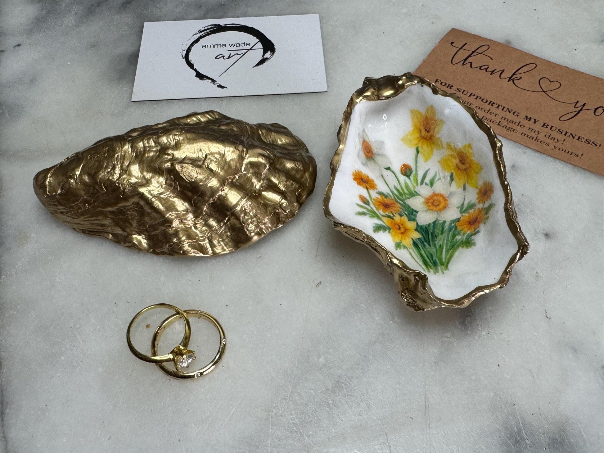 Handmade Dorset oyster shell trinket dish with yellow and orange spring flowers, Daffodils and Marigolds, available in two designs, finished with a gold leaf rim and glossy resin. Features an original Emma Wade Art design created exclusively for this collection. Shown holding rings on a light background, highlighting the natural shell shape and handcrafted coastal style, ideal as a jewellery dish or keepsake gift.