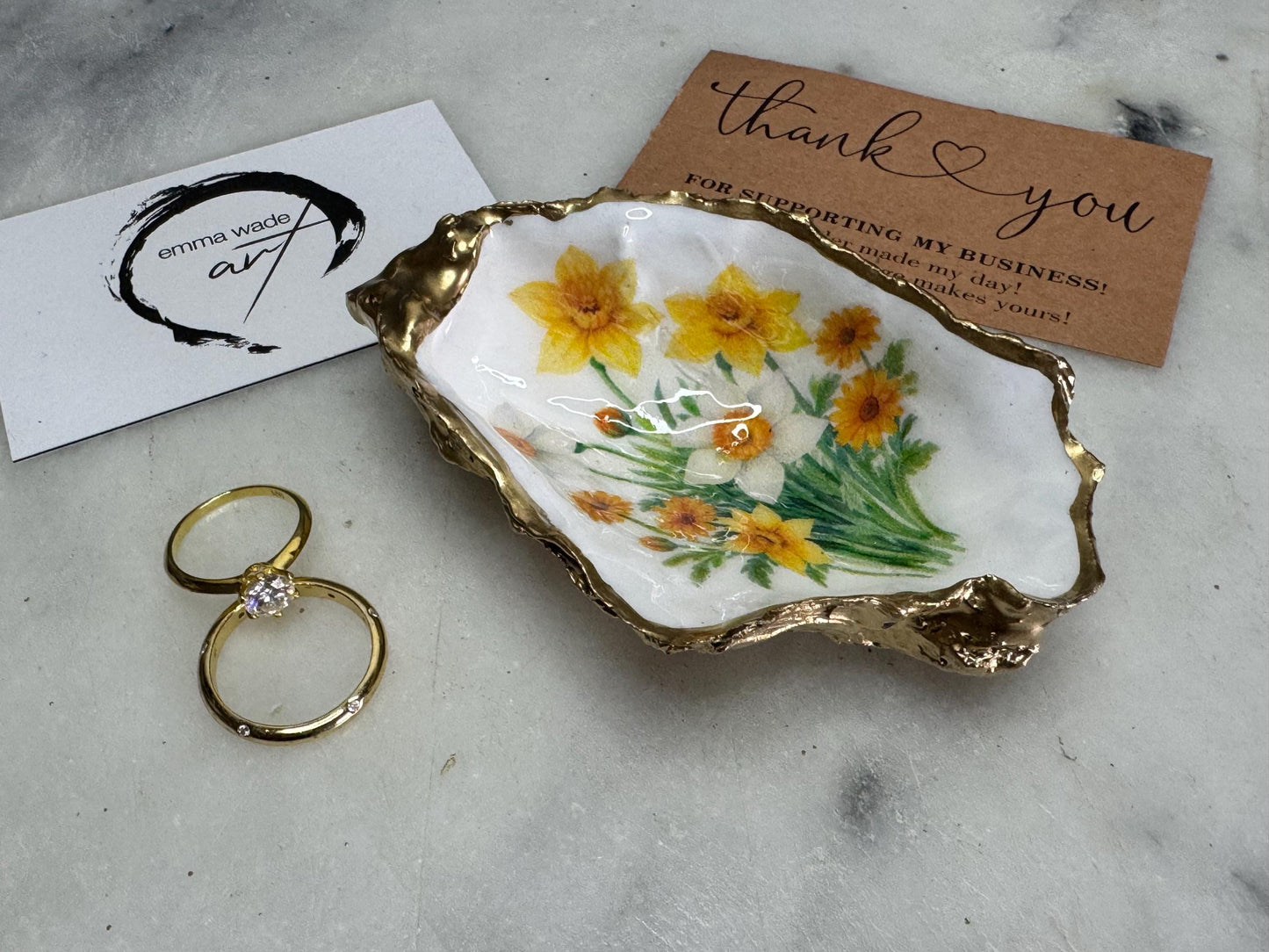 Handmade Dorset oyster shell trinket dish with yellow and orange spring flowers, Daffodils and Marigolds, available in two designs, finished with a gold leaf rim and glossy resin. Features an original Emma Wade Art design created exclusively for this collection. Shown holding rings on a light background, highlighting the natural shell shape and handcrafted coastal style, ideal as a jewellery dish or keepsake gift.