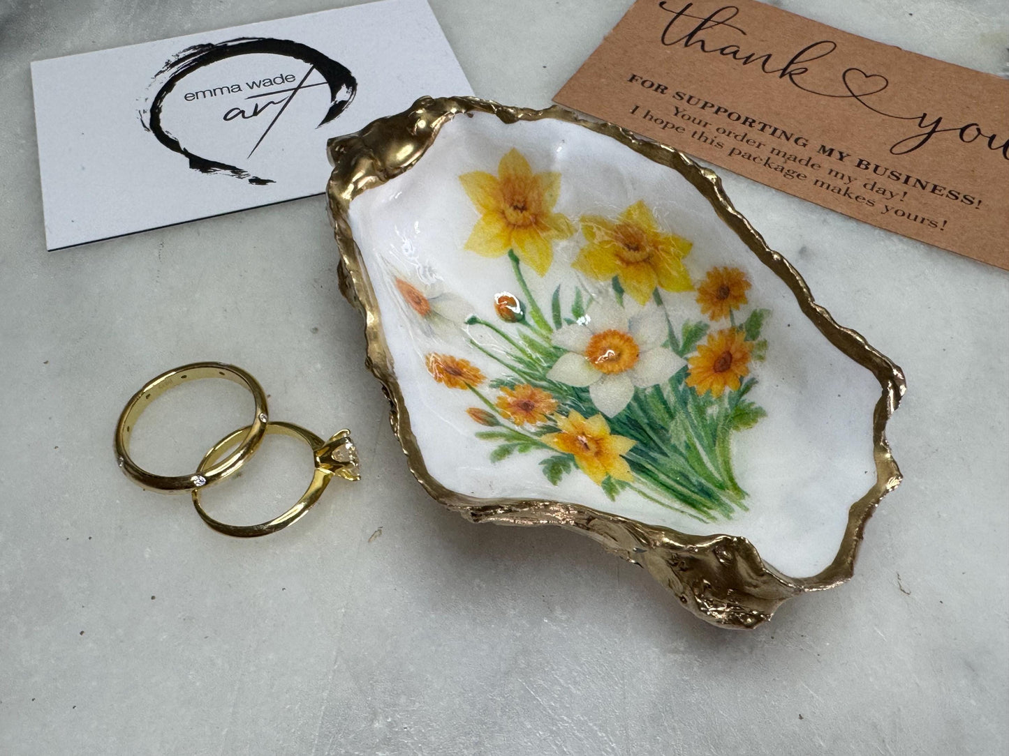 Handmade Dorset oyster shell trinket dish with yellow and orange spring flowers, Daffodils and Marigolds, available in two designs, finished with a gold leaf rim and glossy resin. Features an original Emma Wade Art design created exclusively for this collection. Shown holding rings on a light background, highlighting the natural shell shape and handcrafted coastal style, ideal as a jewellery dish or keepsake gift.
