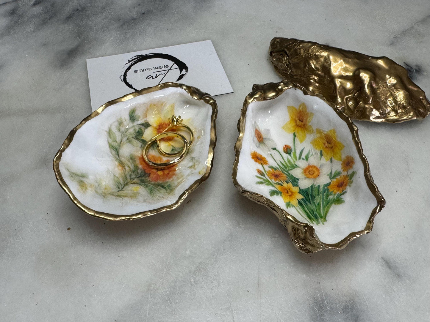 Handmade Dorset oyster shell trinket dish with yellow and orange spring flowers, Daffodils and Marigolds, available in two designs, finished with a gold leaf rim and glossy resin. Features an original Emma Wade Art design created exclusively for this collection. Shown holding rings on a light background, highlighting the natural shell shape and handcrafted coastal style, ideal as a jewellery dish or keepsake gift.