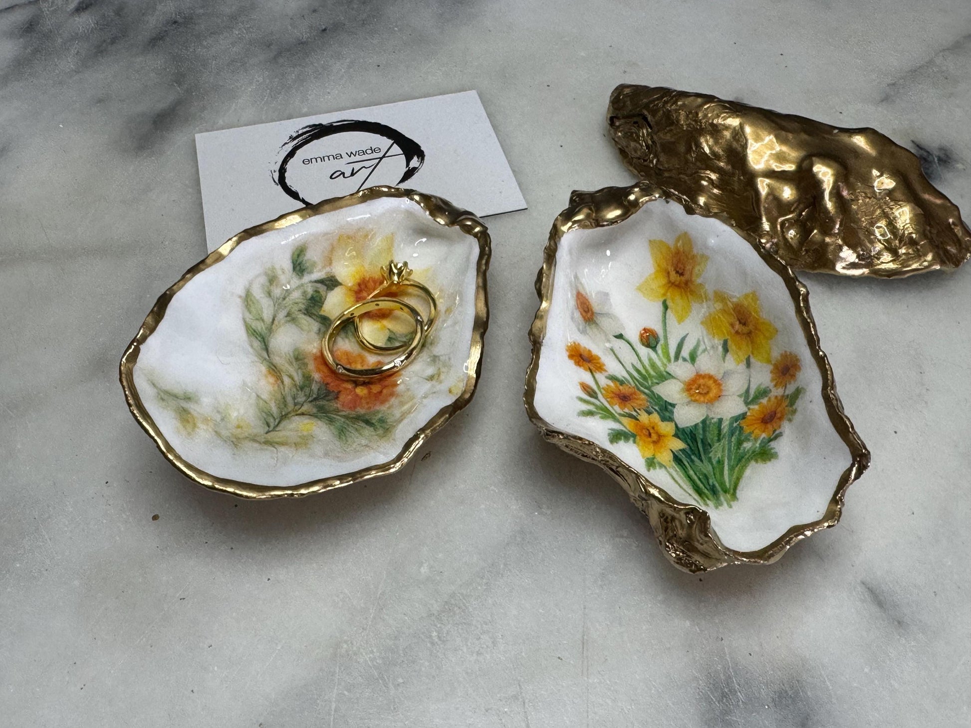 Handmade Dorset oyster shell trinket dish with yellow and orange spring flowers, Daffodils and Marigolds, available in two designs, finished with a gold leaf rim and glossy resin. Features an original Emma Wade Art design created exclusively for this collection. Shown holding rings on a light background, highlighting the natural shell shape and handcrafted coastal style, ideal as a jewellery dish or keepsake gift.