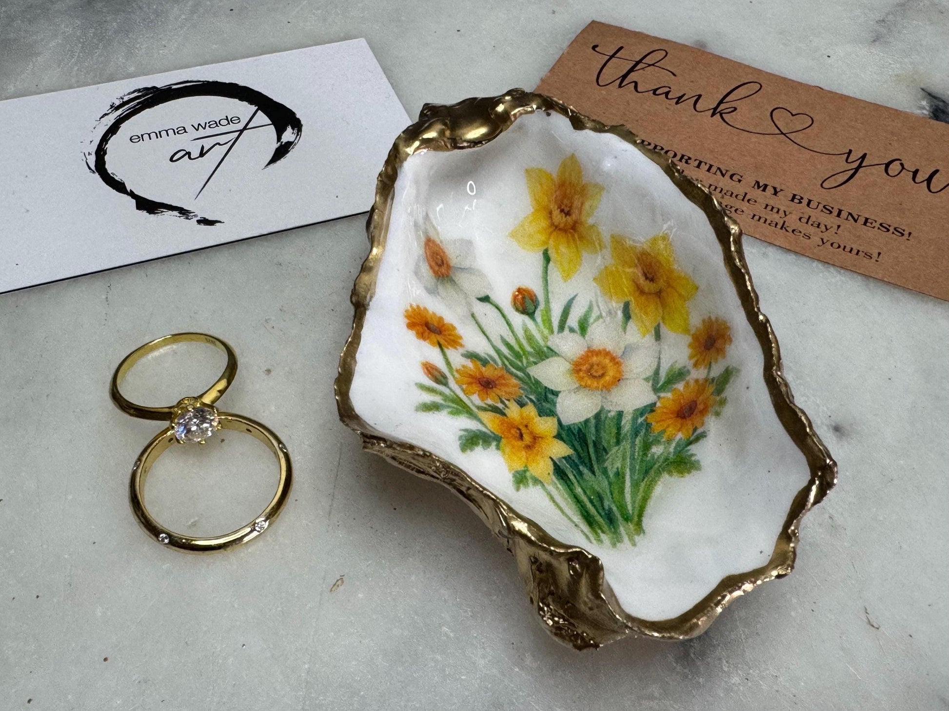 Handmade Dorset oyster shell trinket dish with yellow and orange spring flowers, Daffodils and Marigolds, available in two designs, finished with a gold leaf rim and glossy resin. Features an original Emma Wade Art design created exclusively for this collection. Shown holding rings on a light background, highlighting the natural shell shape and handcrafted coastal style, ideal as a jewellery dish or keepsake gift.