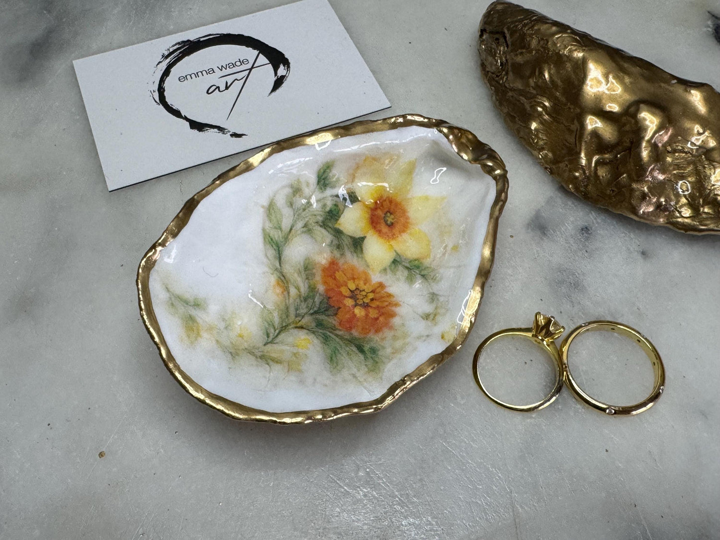 Handmade Dorset oyster shell trinket dish with yellow and orange spring flowers, Daffodils and Marigolds, available in two designs, finished with a gold leaf rim and glossy resin. Features an original Emma Wade Art design created exclusively for this collection. Shown holding rings on a light background, highlighting the natural shell shape and handcrafted coastal style, ideal as a jewellery dish or keepsake gift.