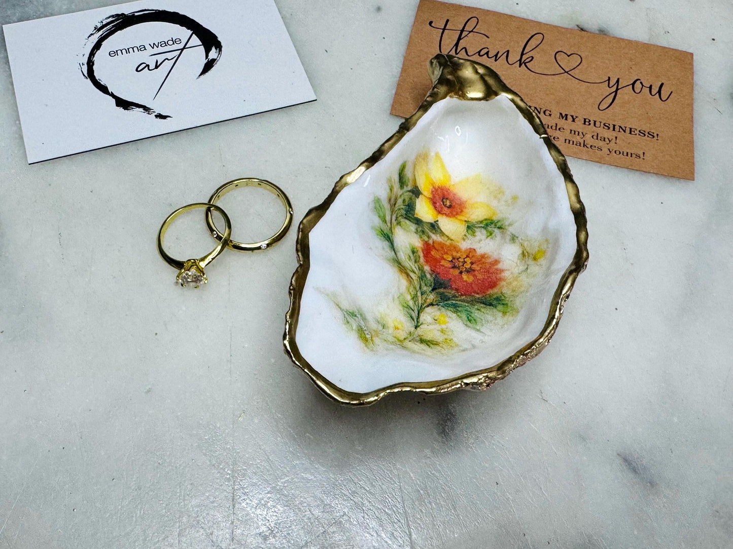 Handmade Dorset oyster shell trinket dish with yellow and orange spring flowers, Daffodils and Marigolds, available in two designs, finished with a gold leaf rim and glossy resin. Features an original Emma Wade Art design created exclusively for this collection. Shown holding rings on a light background, highlighting the natural shell shape and handcrafted coastal style, ideal as a jewellery dish or keepsake gift.