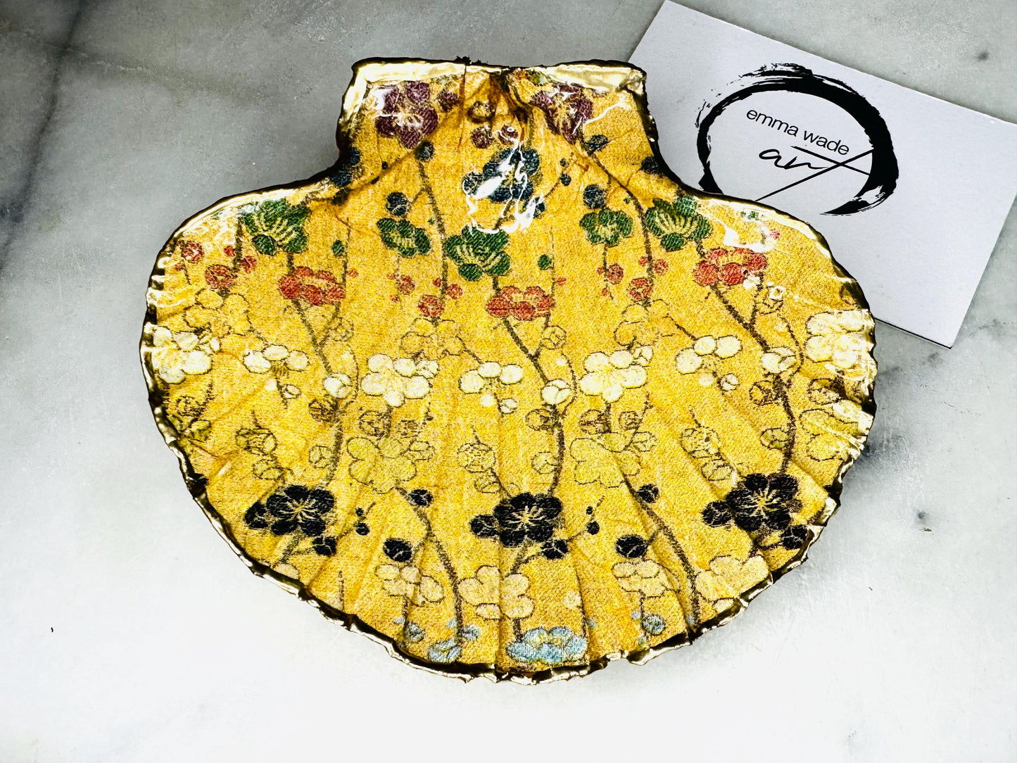 Handcrafted Dorset scallop shell trinket dish featuring a Japanese plum blossom floral design on a rich yellow background, finished with a gold leaf rim and glossy resin. Decorative blossom branches in white, green and black tones, styled on a light marble surface with a gold shell accent. Coastal inspired jewellery tray or ring holder, ideal as a handmade decorative keepsake gift.