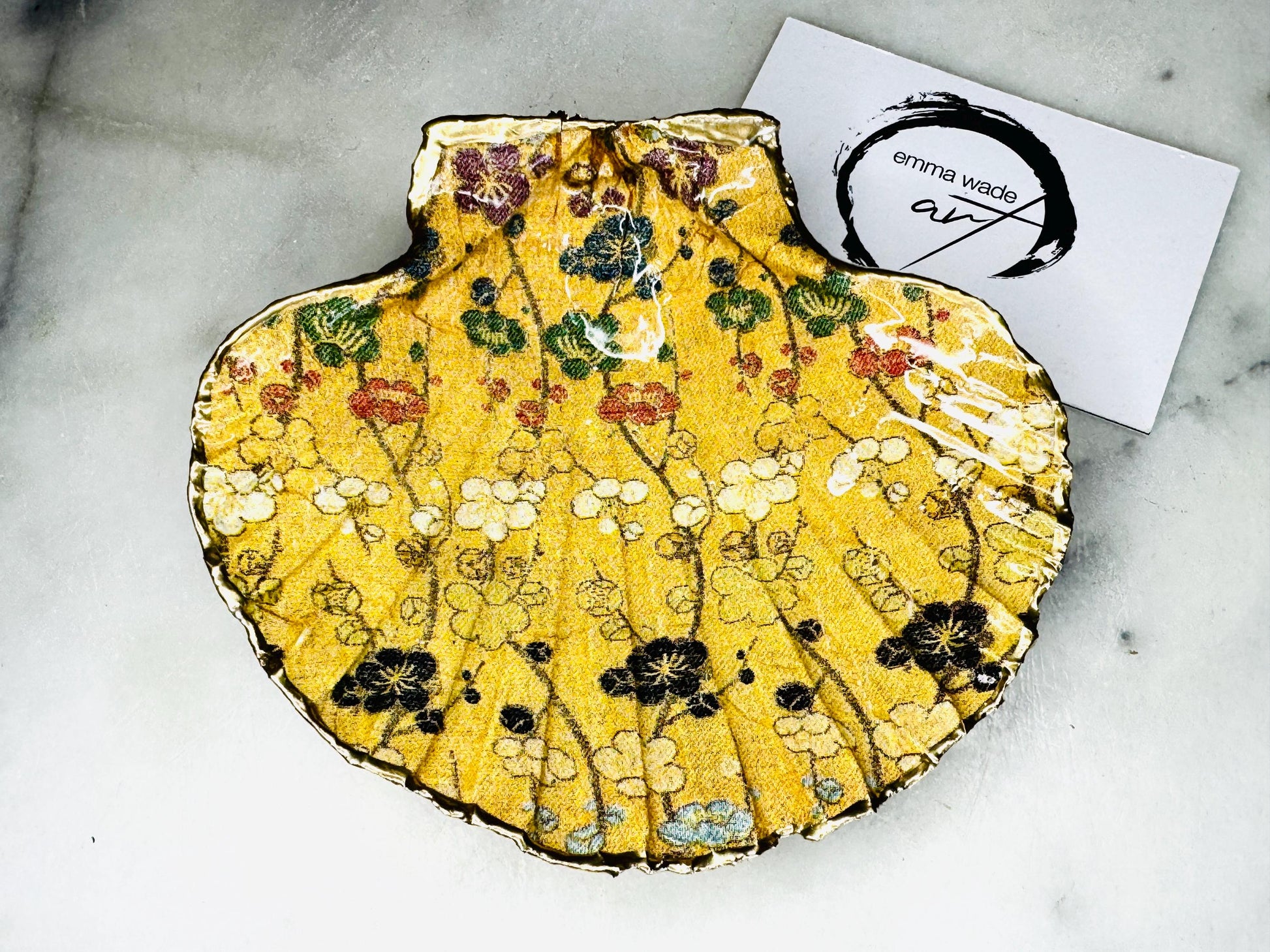 Handcrafted Dorset scallop shell trinket dish featuring a Japanese plum blossom floral design on a rich yellow background, finished with a gold leaf rim and glossy resin. Decorative blossom branches in white, green and black tones, styled on a light marble surface with a gold shell accent. Coastal inspired jewellery tray or ring holder, ideal as a handmade decorative keepsake gift.