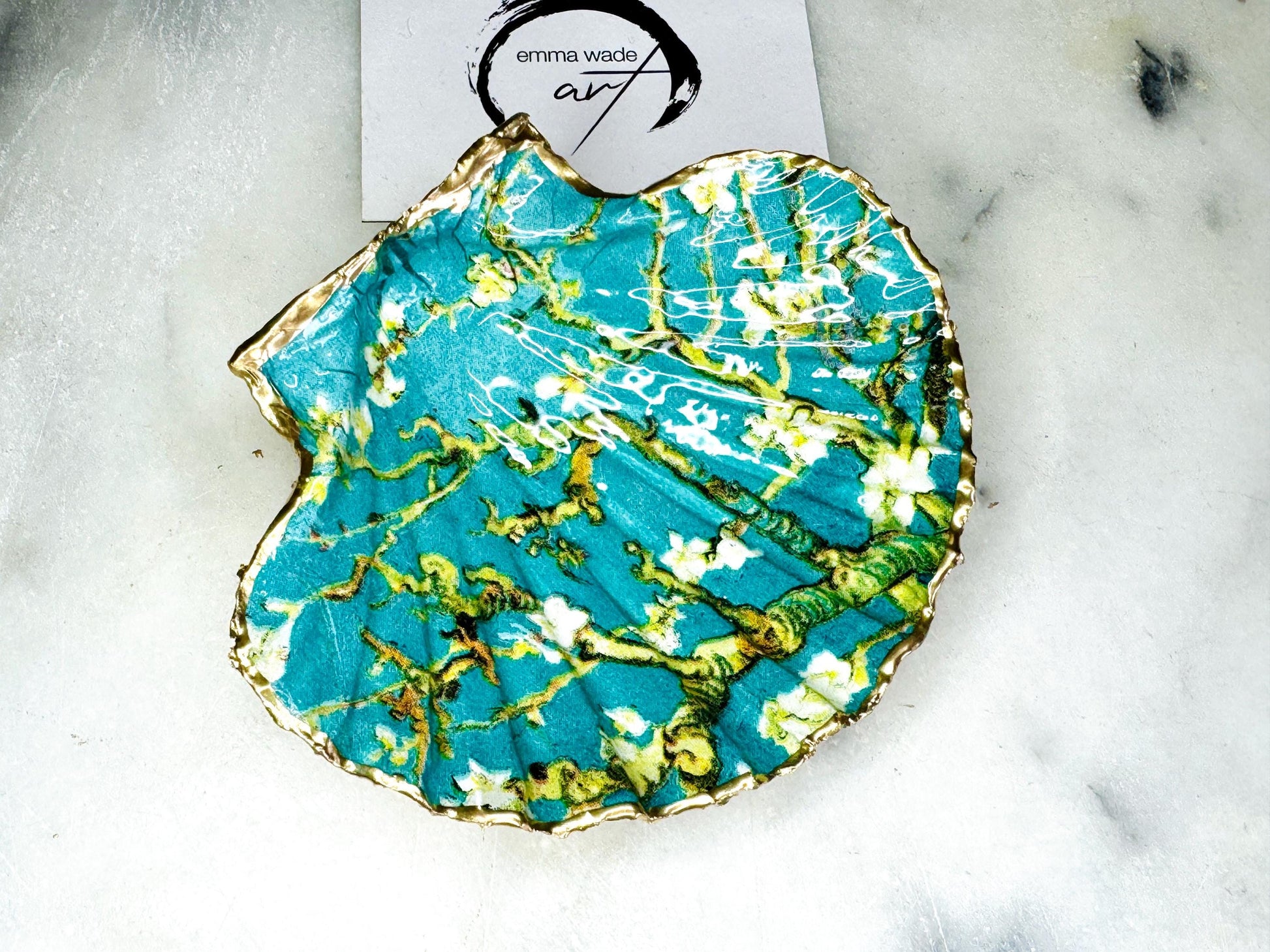 Handcrafted Dorset scallop shell trinket dish inspired by Vincent van Gogh’s Almond Blossom painting. Turquoise blue and green background with branching blossom design and raised gold detailing, finished with liquid gold leaf edging and sealed in high-gloss resin. Decorative shell jewellery holder styled on marble surface, suitable for rings, earrings, trinkets or as a tea light holder. Coastal art-inspired home decor piece.