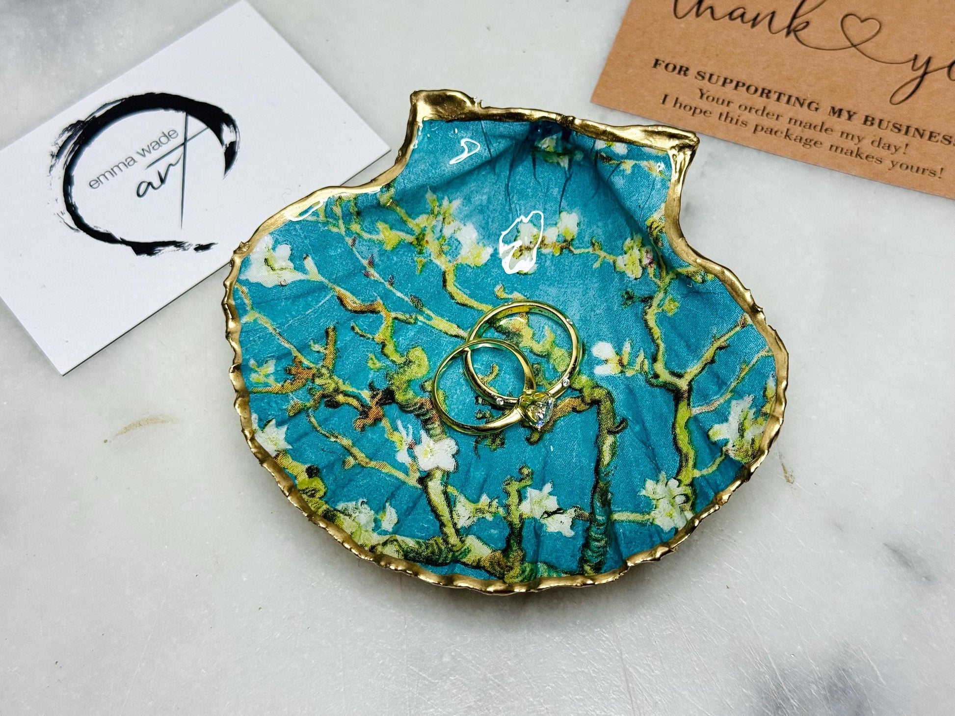 Handcrafted Dorset scallop shell trinket dish inspired by Vincent van Gogh’s Almond Blossom painting. Turquoise blue and green background with branching blossom design and raised gold detailing, finished with liquid gold leaf edging and sealed in high-gloss resin. Decorative shell jewellery holder styled on marble surface, suitable for rings, earrings, trinkets or as a tea light holder. Coastal art-inspired home decor piece.