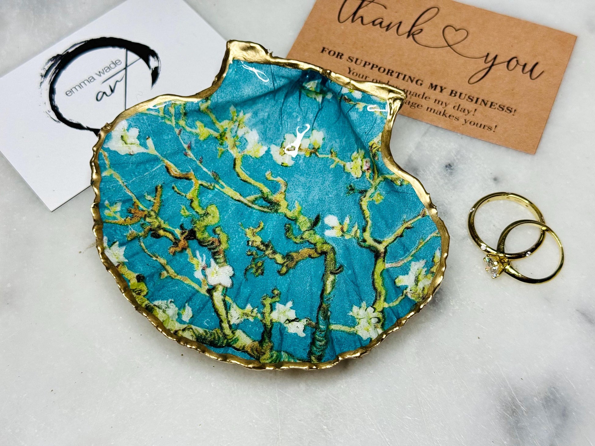 Handcrafted Dorset scallop shell trinket dish inspired by Vincent van Gogh’s Almond Blossom painting. Turquoise blue and green background with branching blossom design and raised gold detailing, finished with liquid gold leaf edging and sealed in high-gloss resin. Decorative shell jewellery holder styled on marble surface, suitable for rings, earrings, trinkets or as a tea light holder. Coastal art-inspired home decor piece.