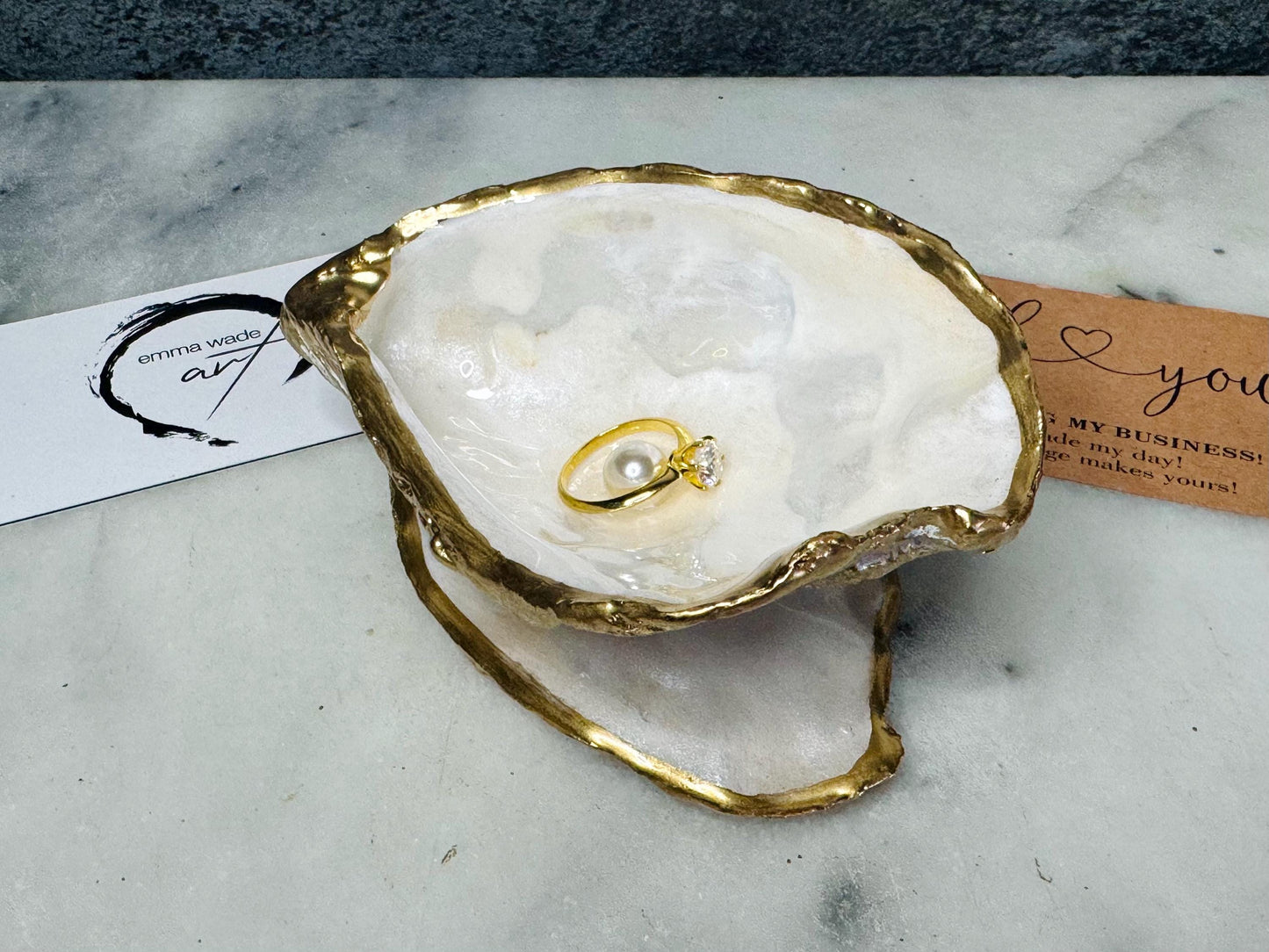 Handcrafted Dorset oyster shell ring box with matching natural shell lid, featuring a luminous inset Swarovski pearl nestled inside the cupped shell. Interior has a soft pearlescent white sheen and is sealed with crystal-clear resin. Optional outer finish in natural pearl tone or pearl with subtle gold leaf highlights. Coastal-inspired engagement proposal ring holder and wedding keepsake, styled on marble surface.