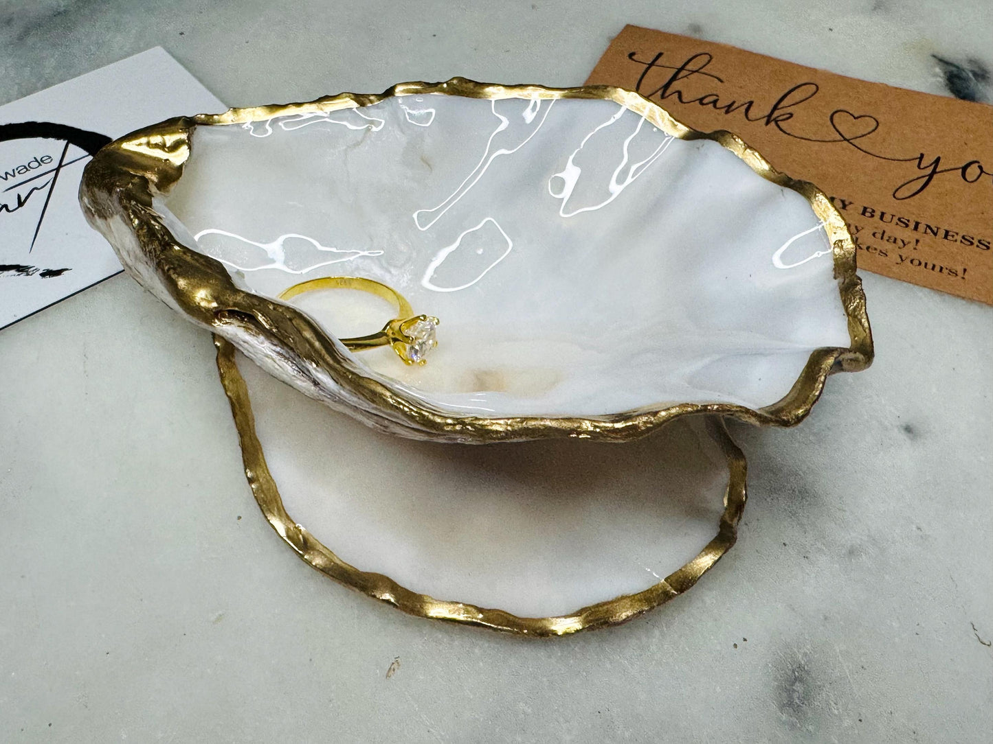 Handcrafted Dorset oyster shell ring box with matching natural shell lid, featuring a luminous inset Swarovski pearl nestled inside the cupped shell. Interior has a soft pearlescent white sheen and is sealed with crystal-clear resin. Optional outer finish in natural pearl tone or pearl with subtle gold leaf highlights. Coastal-inspired engagement proposal ring holder and wedding keepsake, styled on marble surface.