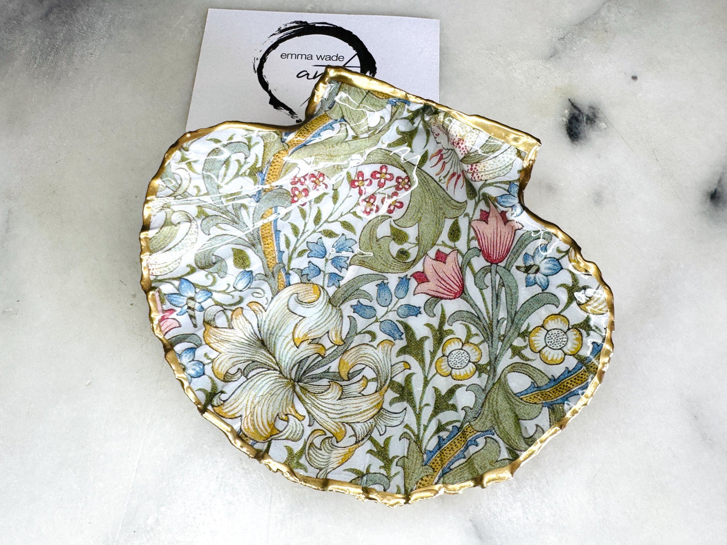 Handcrafted Dorset scallop shell trinket dish featuring William Morris Golden Lily floral design. Natural shell edged with hand applied gold leaf and sealed with glossy resin. Heritage floral jewellery holder displayed with rings on marble surface. Elegant coastal home decor gift.