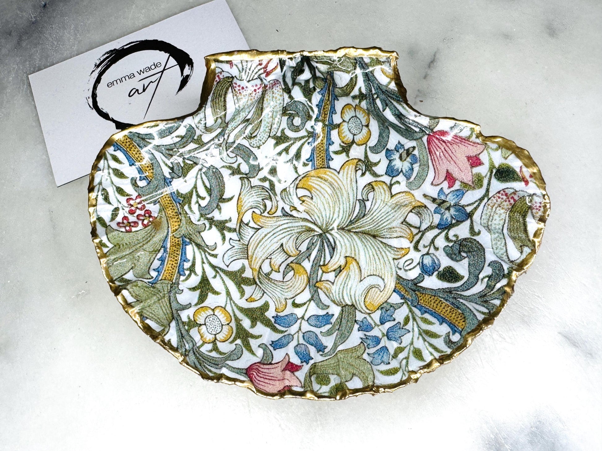 Handcrafted Dorset scallop shell trinket dish featuring William Morris Golden Lily floral design. Natural shell edged with hand applied gold leaf and sealed with glossy resin. Heritage floral jewellery holder displayed with rings on marble surface. Elegant coastal home decor gift.