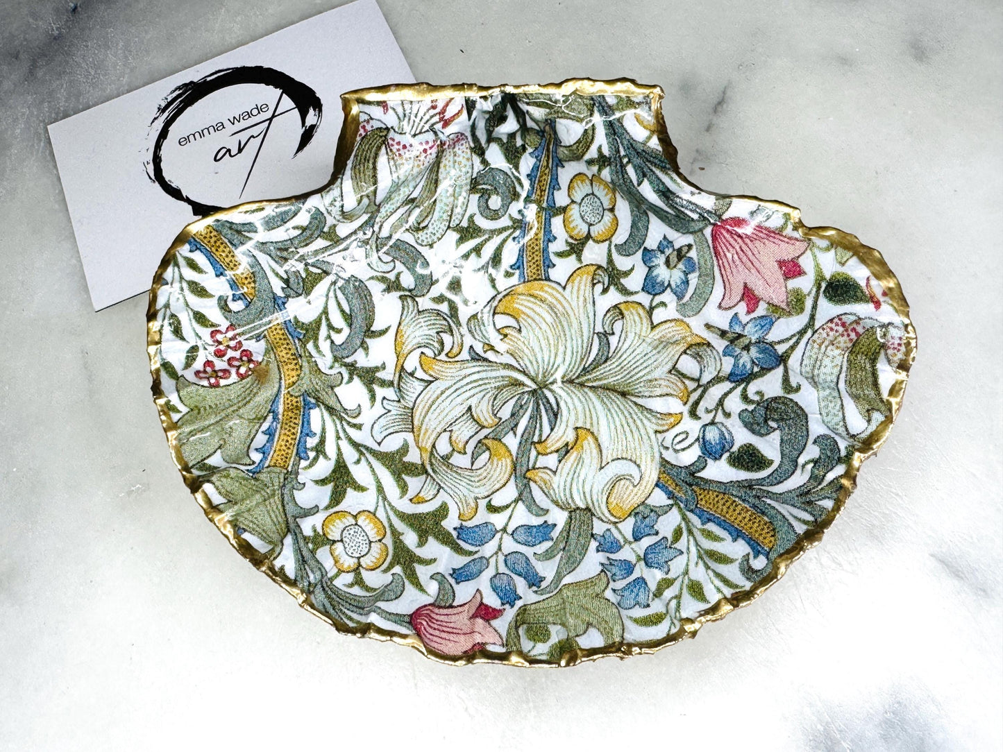 Handcrafted Dorset scallop shell trinket dish featuring William Morris Golden Lily floral design. Natural shell edged with hand applied gold leaf and sealed with glossy resin. Heritage floral jewellery holder displayed with rings on marble surface. Elegant coastal home decor gift.