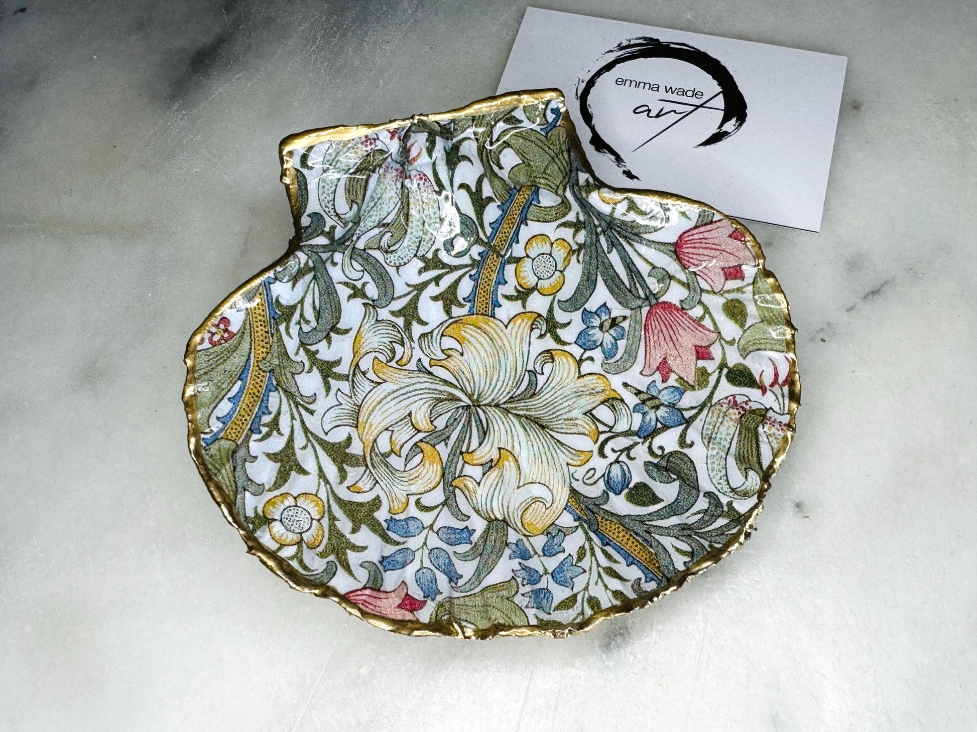 Handcrafted Dorset scallop shell trinket dish featuring William Morris Golden Lily floral design. Natural shell edged with hand applied gold leaf and sealed with glossy resin. Heritage floral jewellery holder displayed with rings on marble surface. Elegant coastal home decor gift.