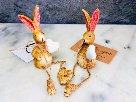 Hand finished rustic bunny shelf sitters holding white wooden hearts, gold and grey rabbit ornaments with pink ears and dangling jute legs, styled on marble surface as charming year-round home decor.