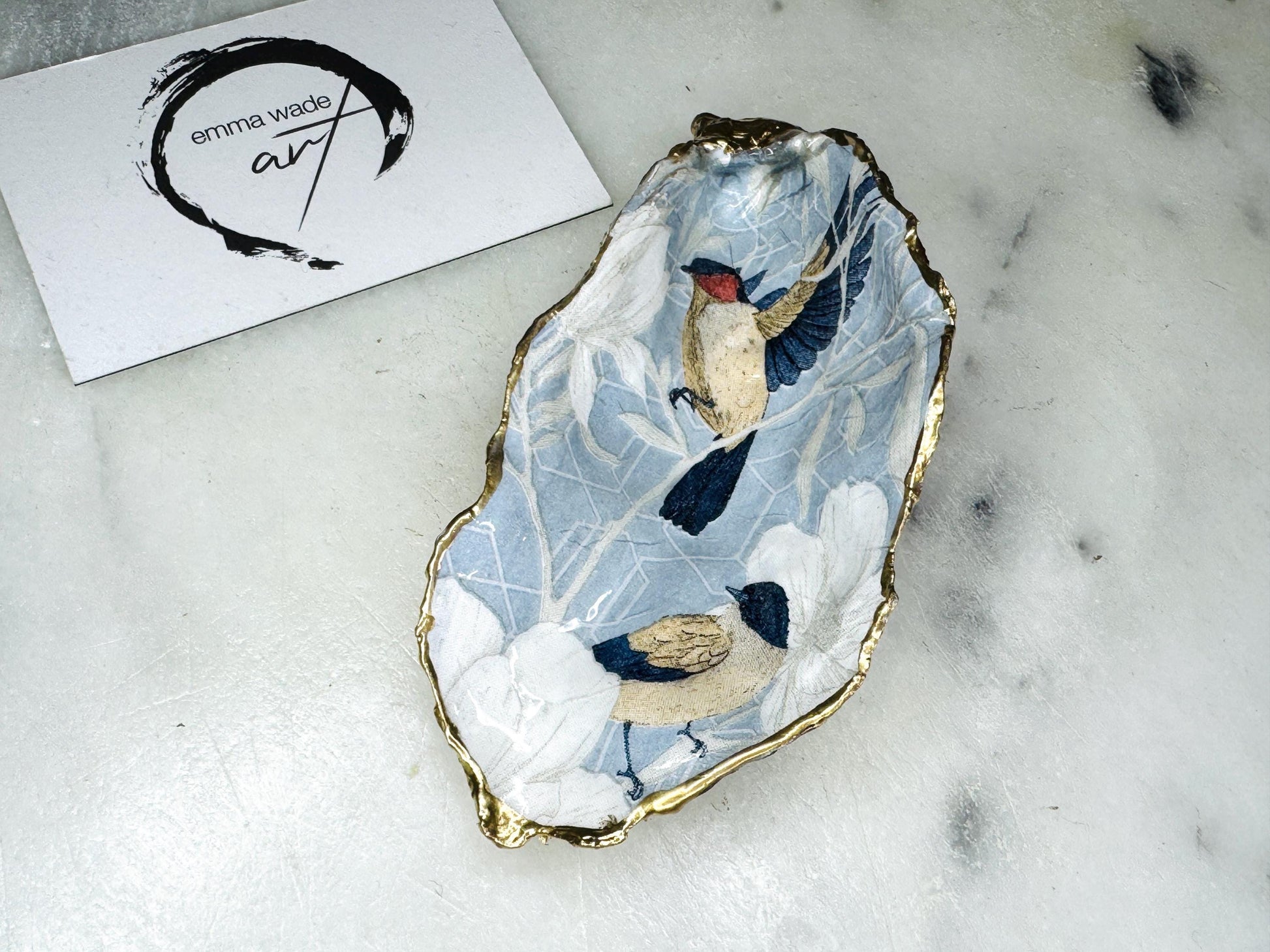 Handmade Dorset oyster shell trinket dish featuring elegant blue bird design with hand gilded gold leaf edging and glossy crystal clear resin finish, luxury coastal jewellery holder and decorative ring dish inspired by birds of paradise and coastal nature