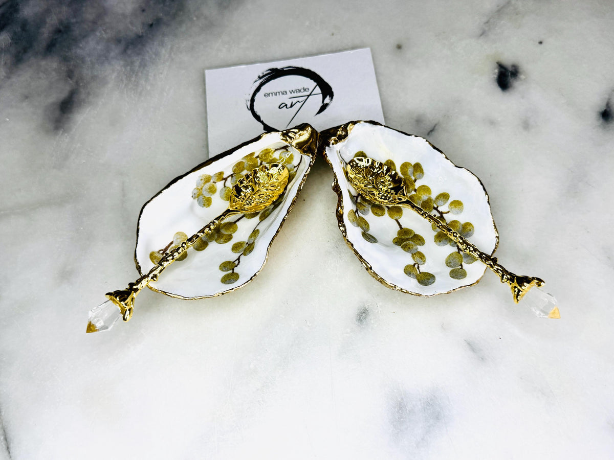 Pair of white and gold oyster shell salt and pepper pinch pots with delicate berry botanical design, hand gilded gold leaf edging and glossy resin finish. Handmade Dorset coastal shell dishes styled with gold crystal spoons on a marble background.