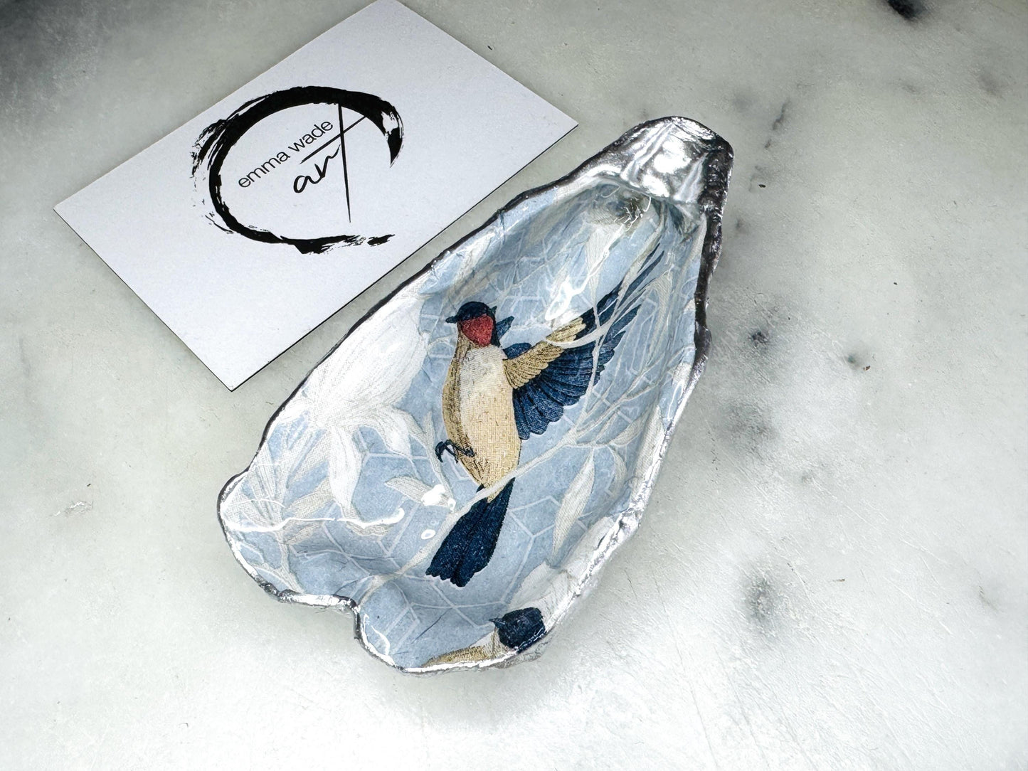 Handmade Dorset oyster shell trinket dish featuring elegant blue bird design with hand gilded gold leaf edging and glossy crystal clear resin finish, luxury coastal jewellery holder and decorative ring dish inspired by birds of paradise and coastal nature