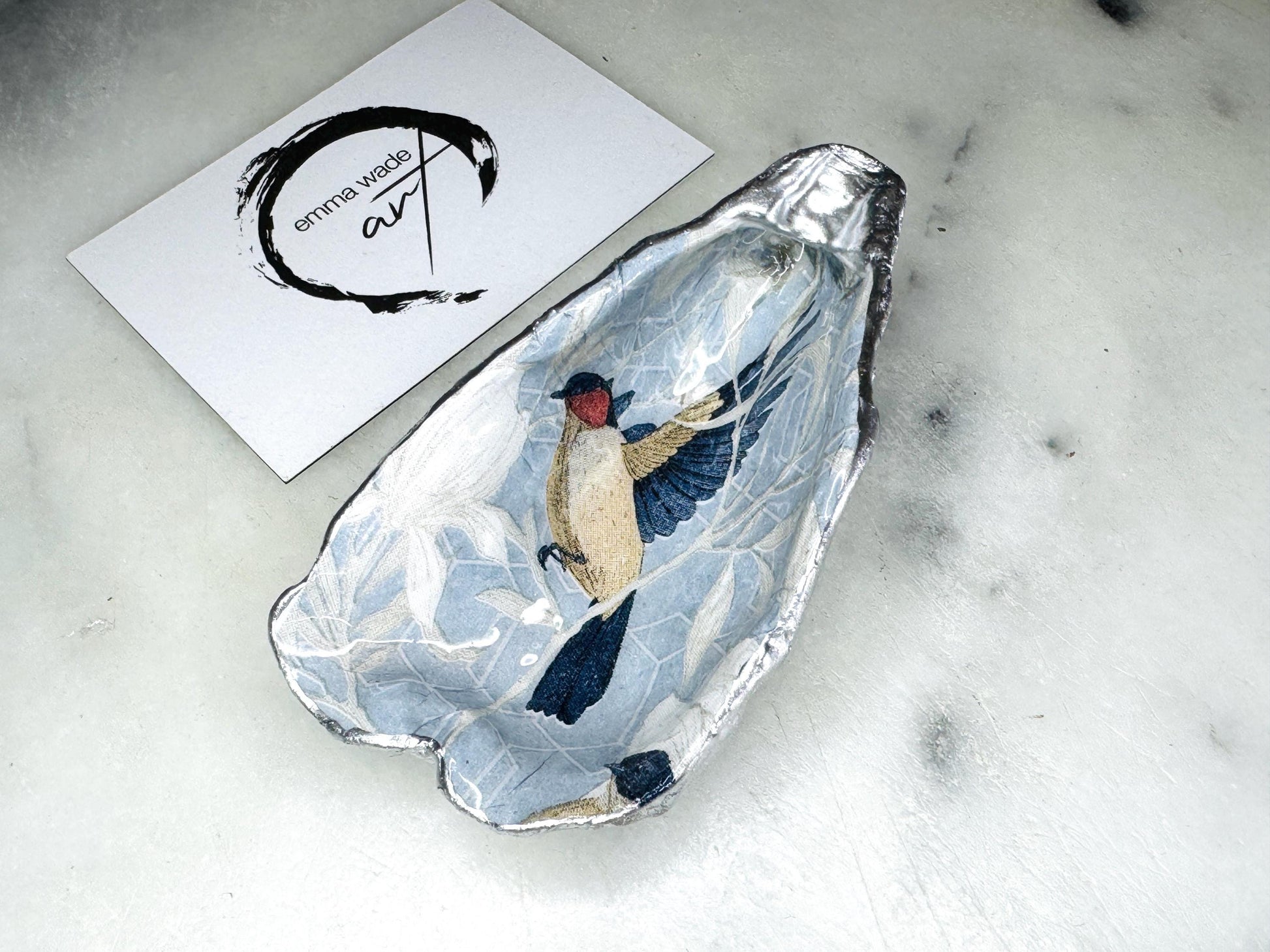 Handmade Dorset oyster shell trinket dish featuring elegant blue bird design with hand gilded gold leaf edging and glossy crystal clear resin finish, luxury coastal jewellery holder and decorative ring dish inspired by birds of paradise and coastal nature