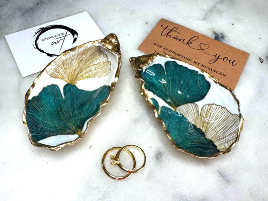 Handmade Dorset oyster shell ring dish featuring Japanese Ginkgo floral leaf artwork in emerald green, gold, and pearl tones with hand-applied liquid gold leaf edging. Luxury coastal jewellery tray sealed in glossy crystal-clear epoxy resin, styled with gold rings.