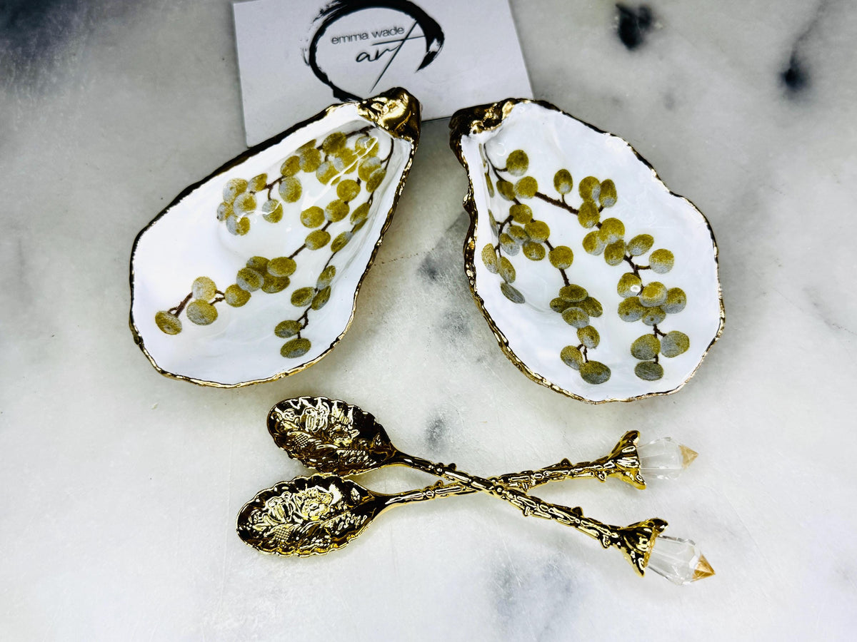 Pair of white and gold oyster shell salt and pepper pinch pots with delicate berry botanical design, hand gilded gold leaf edging and glossy resin finish. Handmade Dorset coastal shell dishes styled with gold crystal spoons on a marble background.