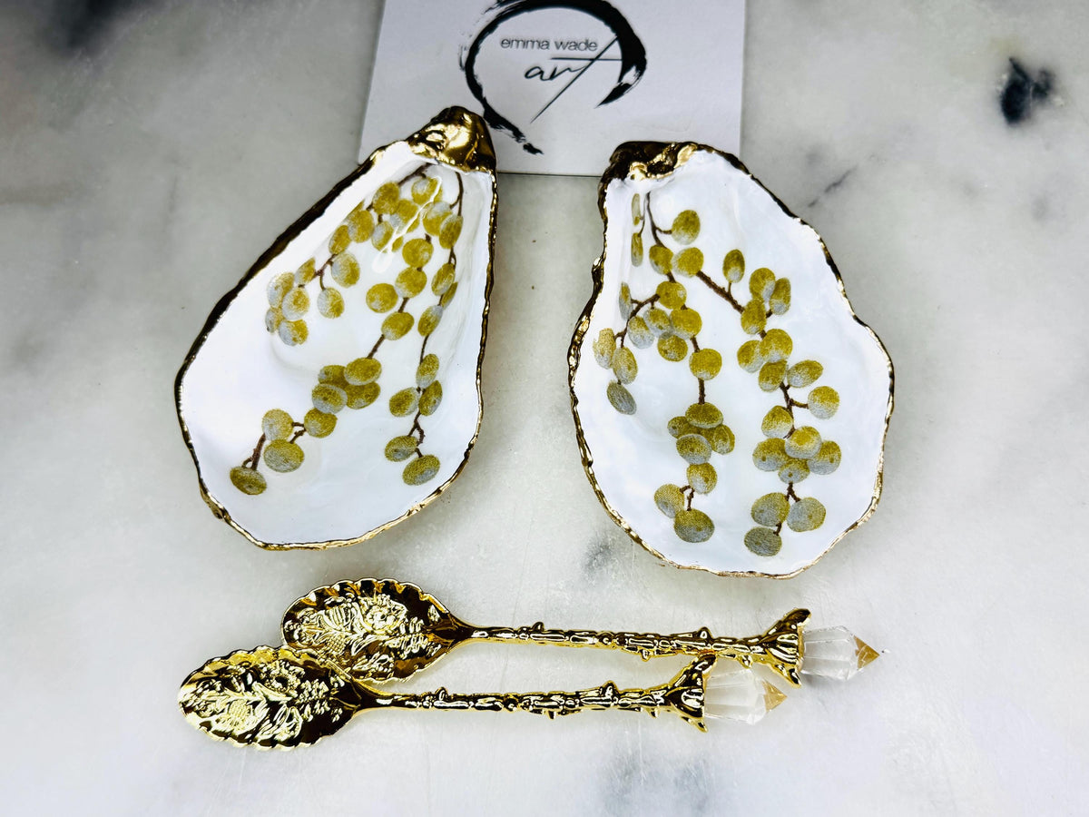 Pair of white and gold oyster shell salt and pepper pinch pots with delicate berry botanical design, hand gilded gold leaf edging and glossy resin finish. Handmade Dorset coastal shell dishes styled with gold crystal spoons on a marble background.